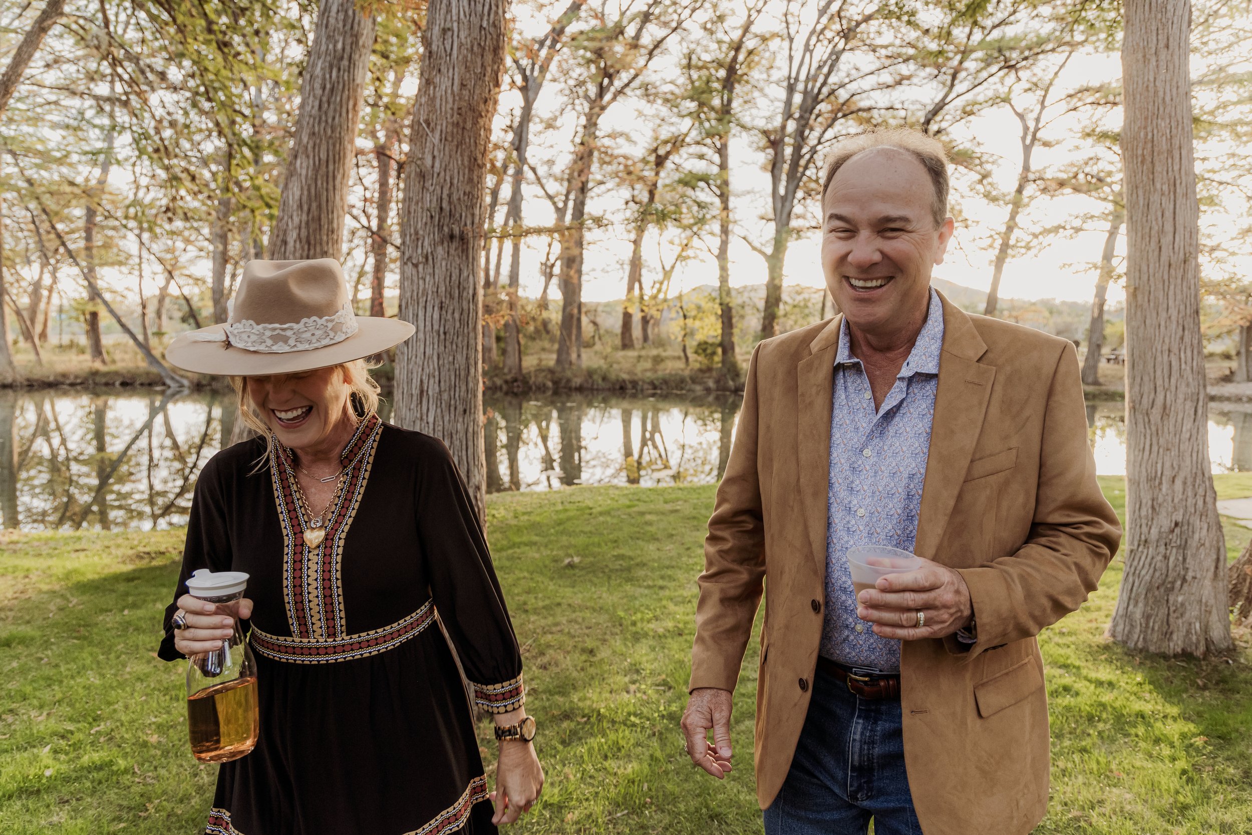 Two people, a woman and a man, laughing and enjoying a drink outdoors near a pond surrounded by trees in warm late afternoon light.