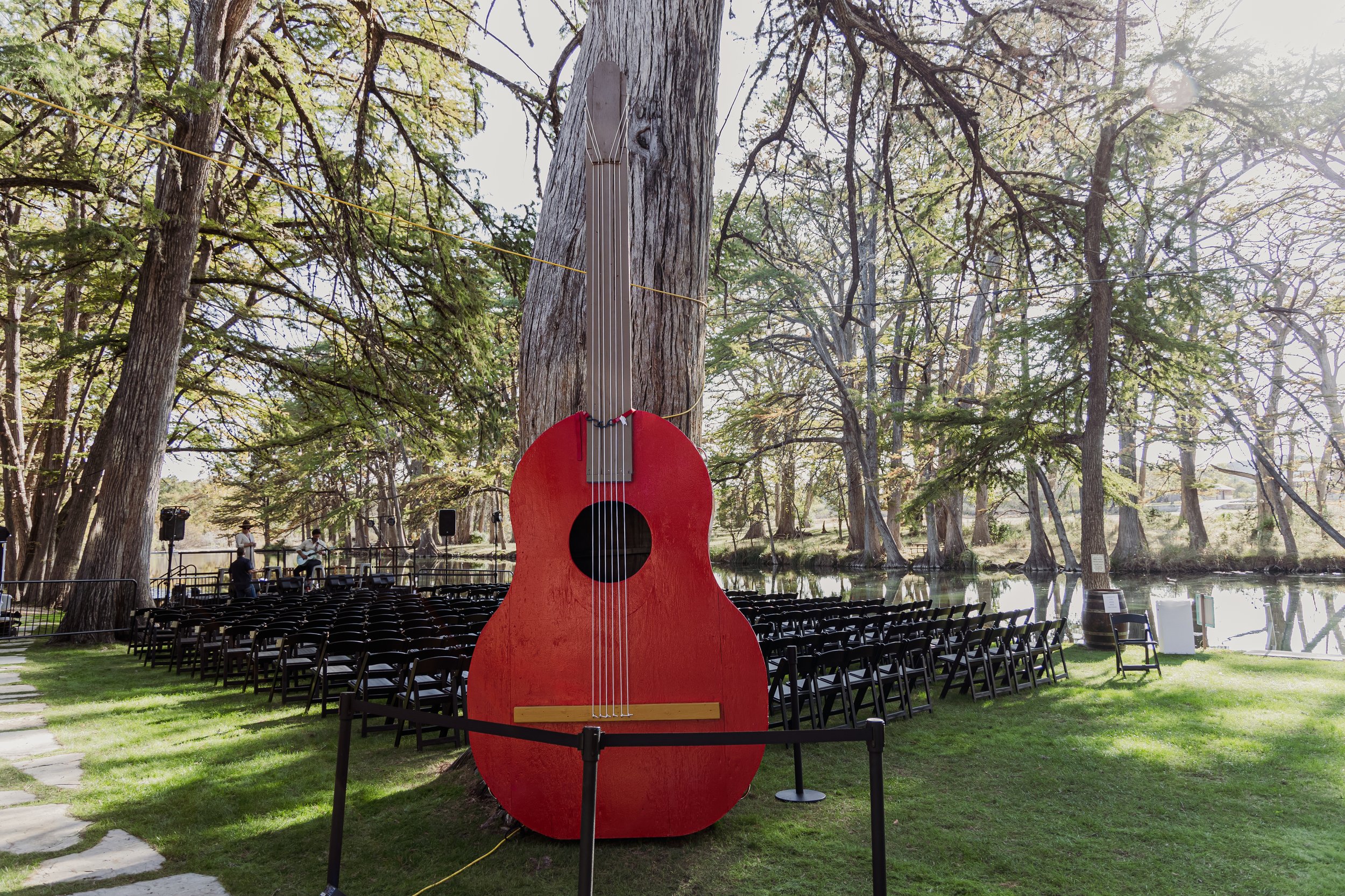Large red guitar sculpture outdoors near a pond with black chairs arranged for an event, surrounded by trees, with a stage and musicians in the background.