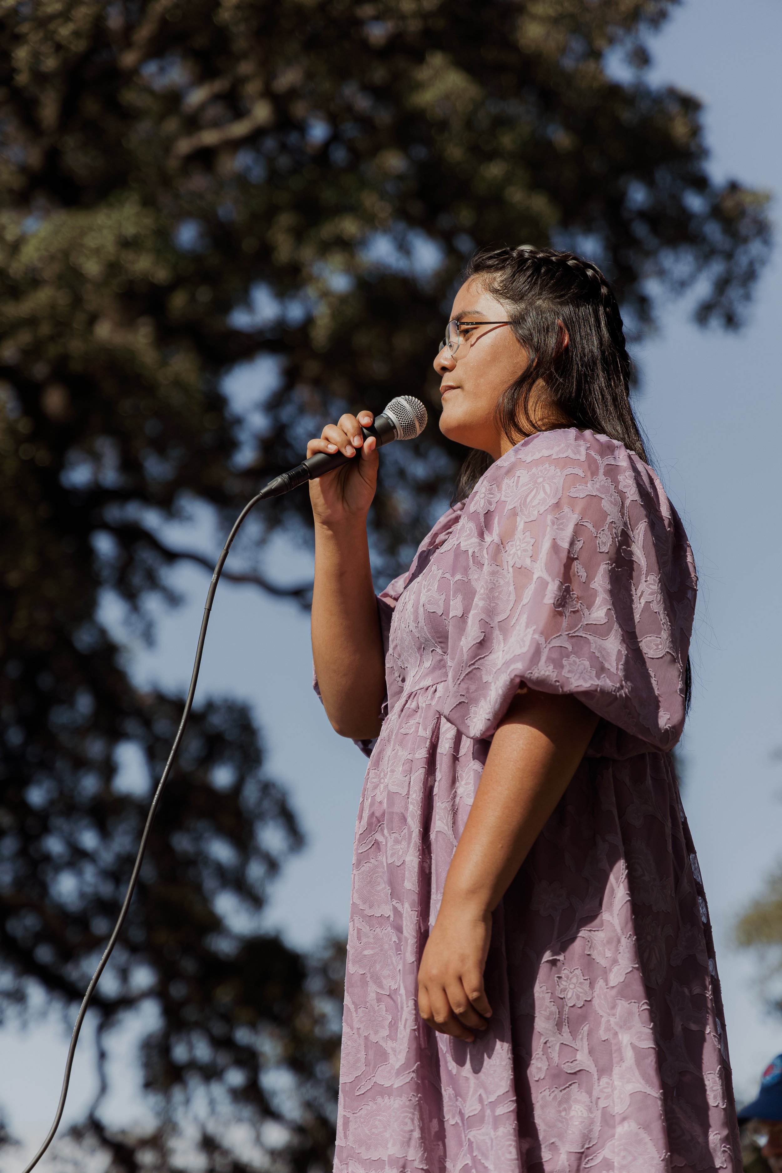 A woman with glasses and long black hair, wearing a lavender dress, standing outdoors, holding a microphone, with trees and a blue sky in the background.
