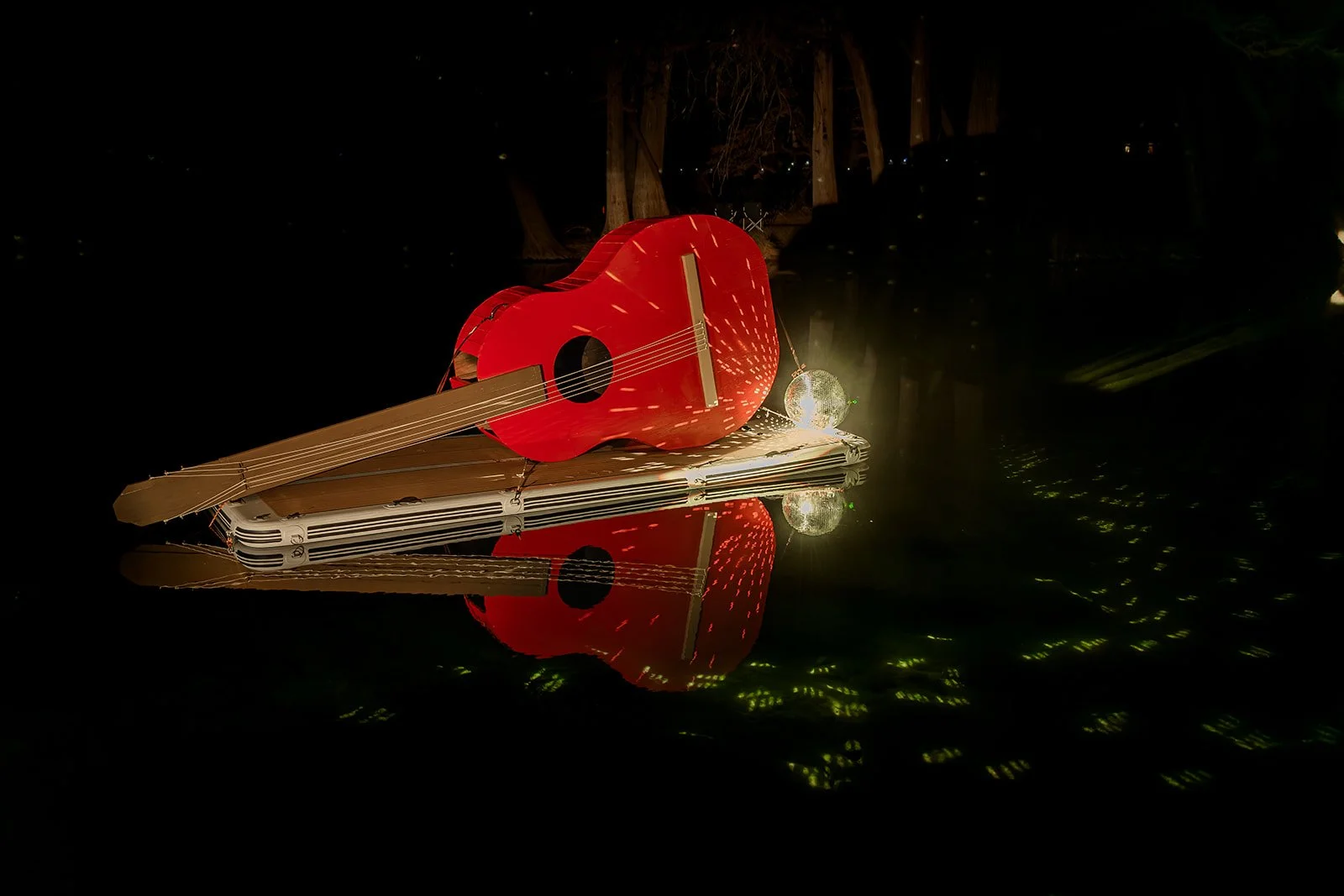 Nighttime scene with illuminated cardboard guitar, small disco ball, lantern, and reflections on water.