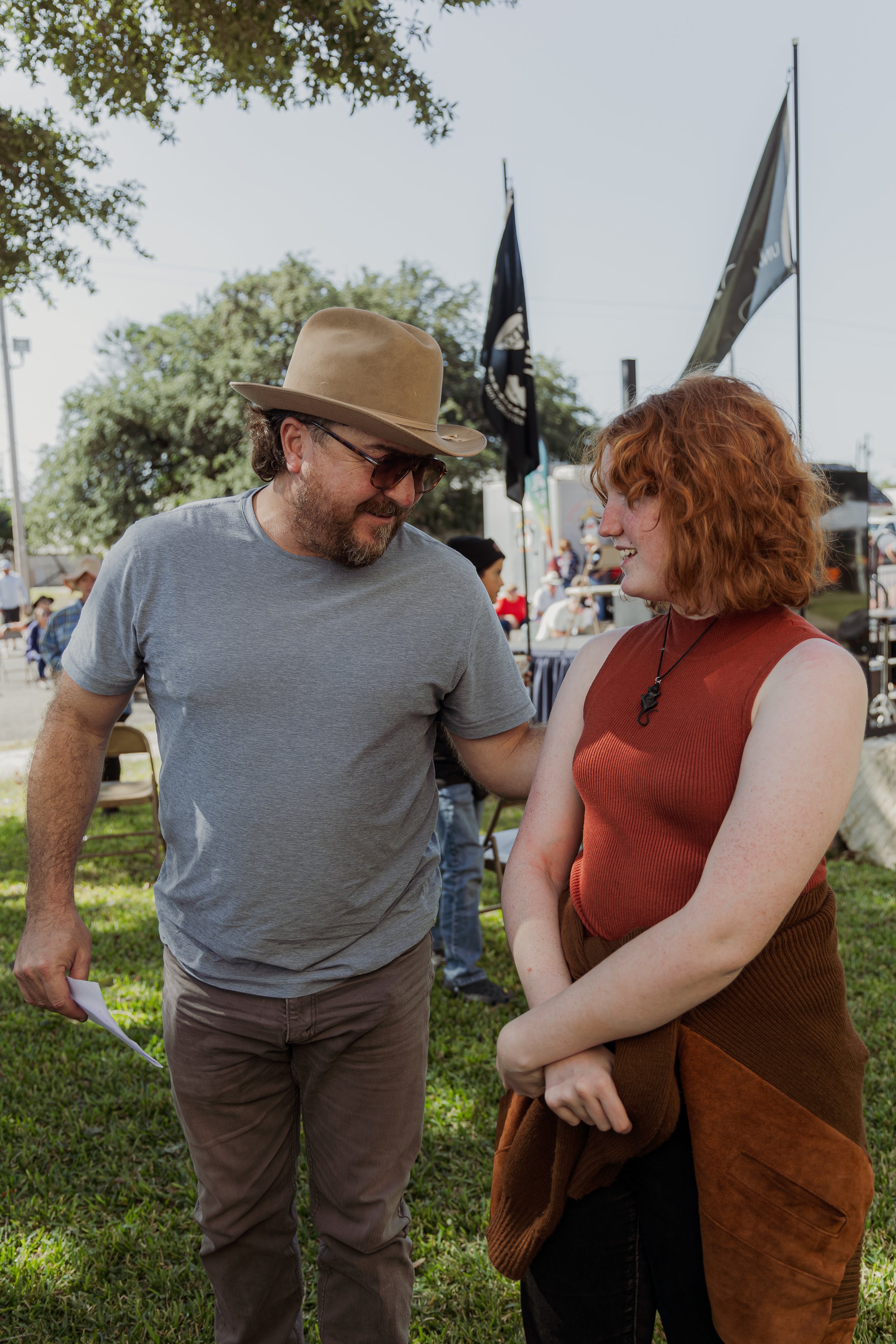A man with a beige hat, glasses, and a gray t-shirt is smiling and talking to a red-haired woman wearing a sleeveless rust-colored top. They are outdoors at a festival or fair with flags and booths in the background.