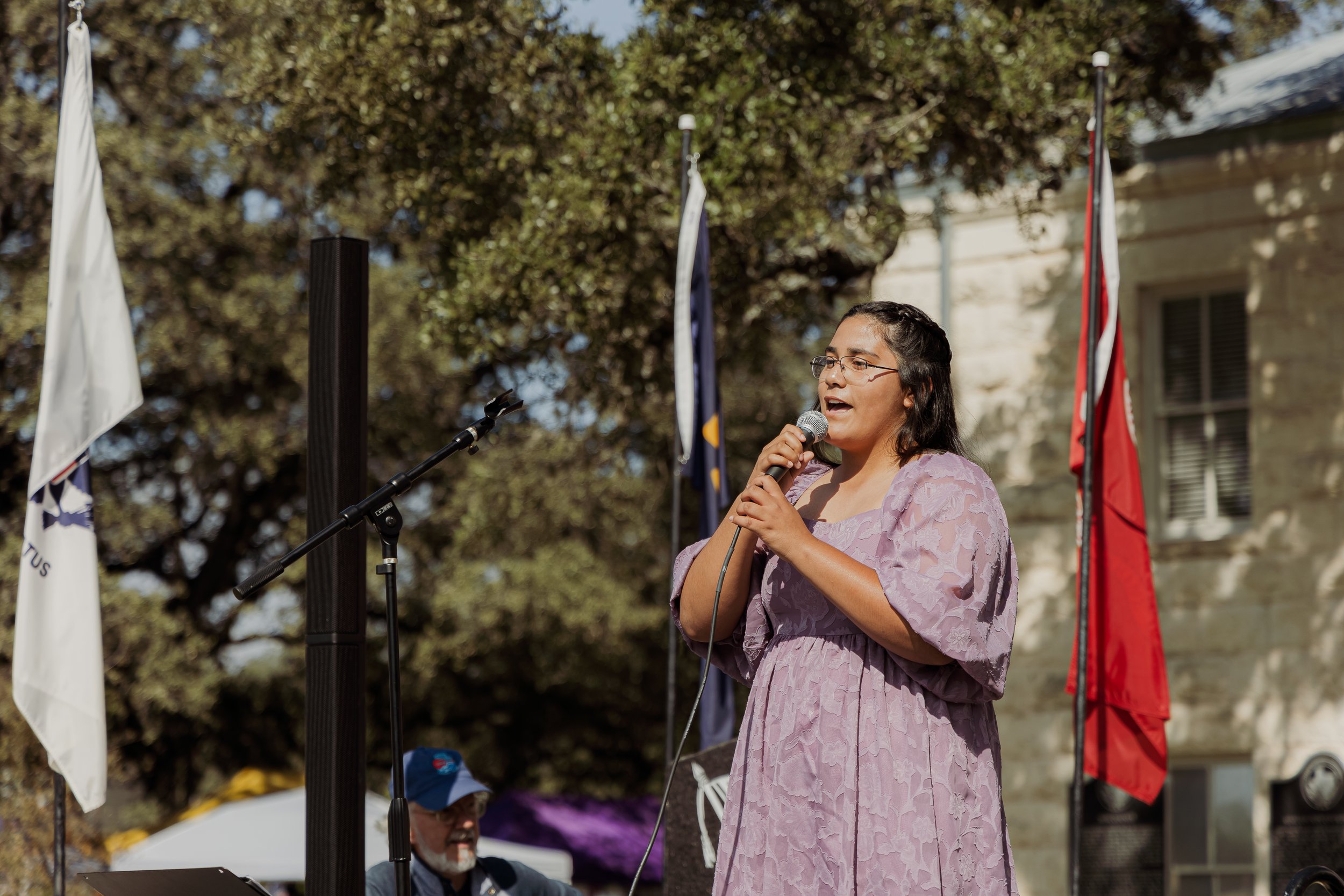 A woman in a light purple dress speaking into a microphone at an outdoor event, with flags and a crowd in the background.