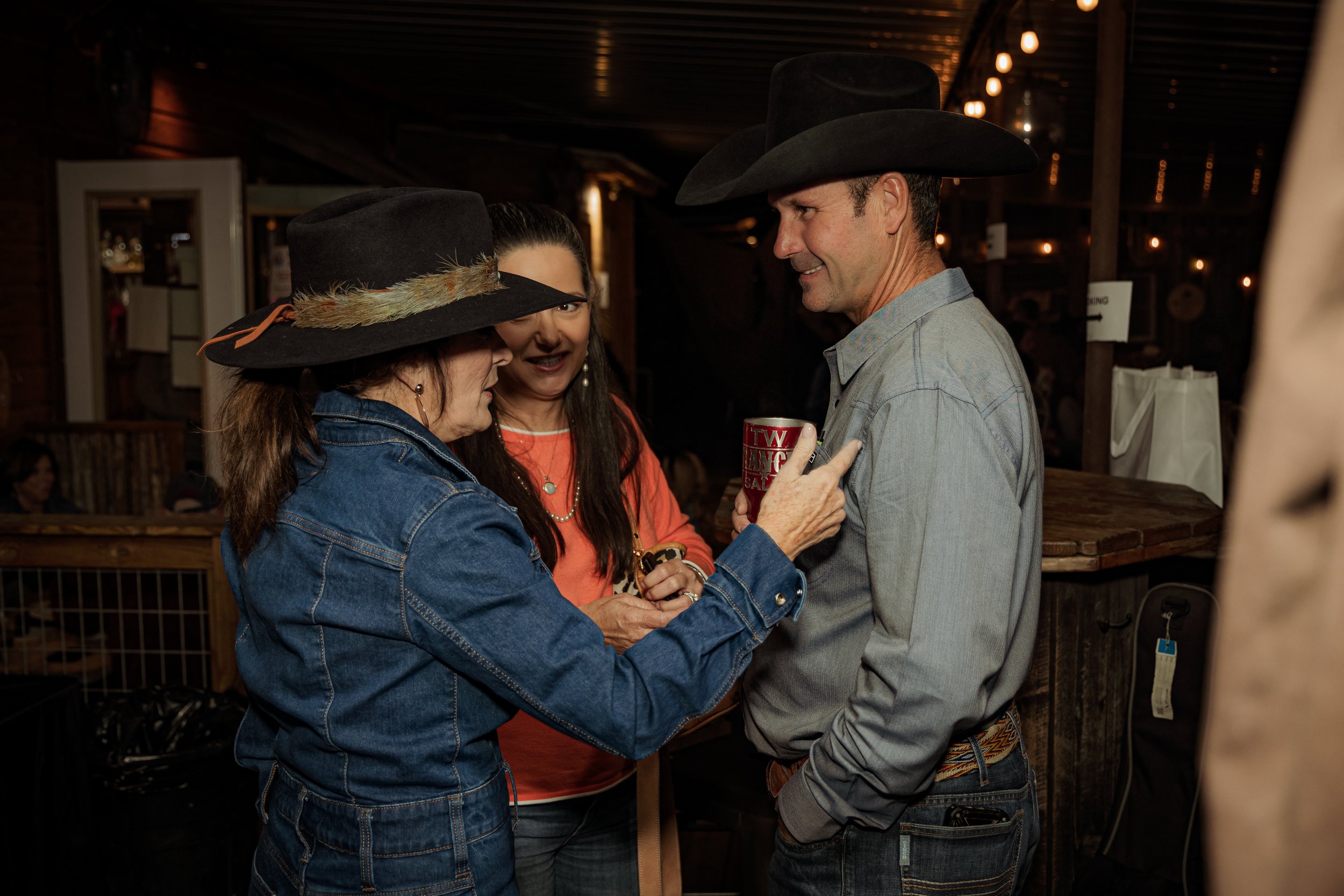 Three people wearing cowboy hats chat and smile inside a dimly lit rustic bar or restaurant, with one person holding a decorated red mug.