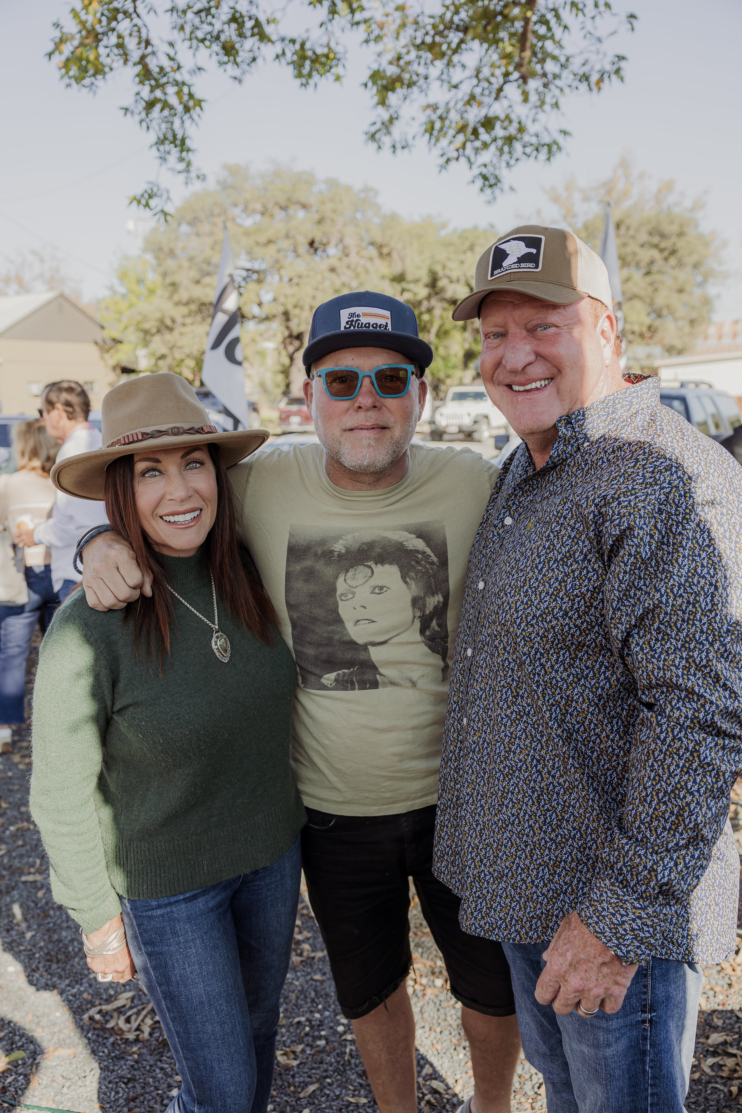 Three people standing outdoors under a tree, smiling, with cars and other people in the background. They are dressed casually, with the woman on the left wearing a hat and green sweater, the man in the middle wearing sunglasses and a graphic t-shirt,