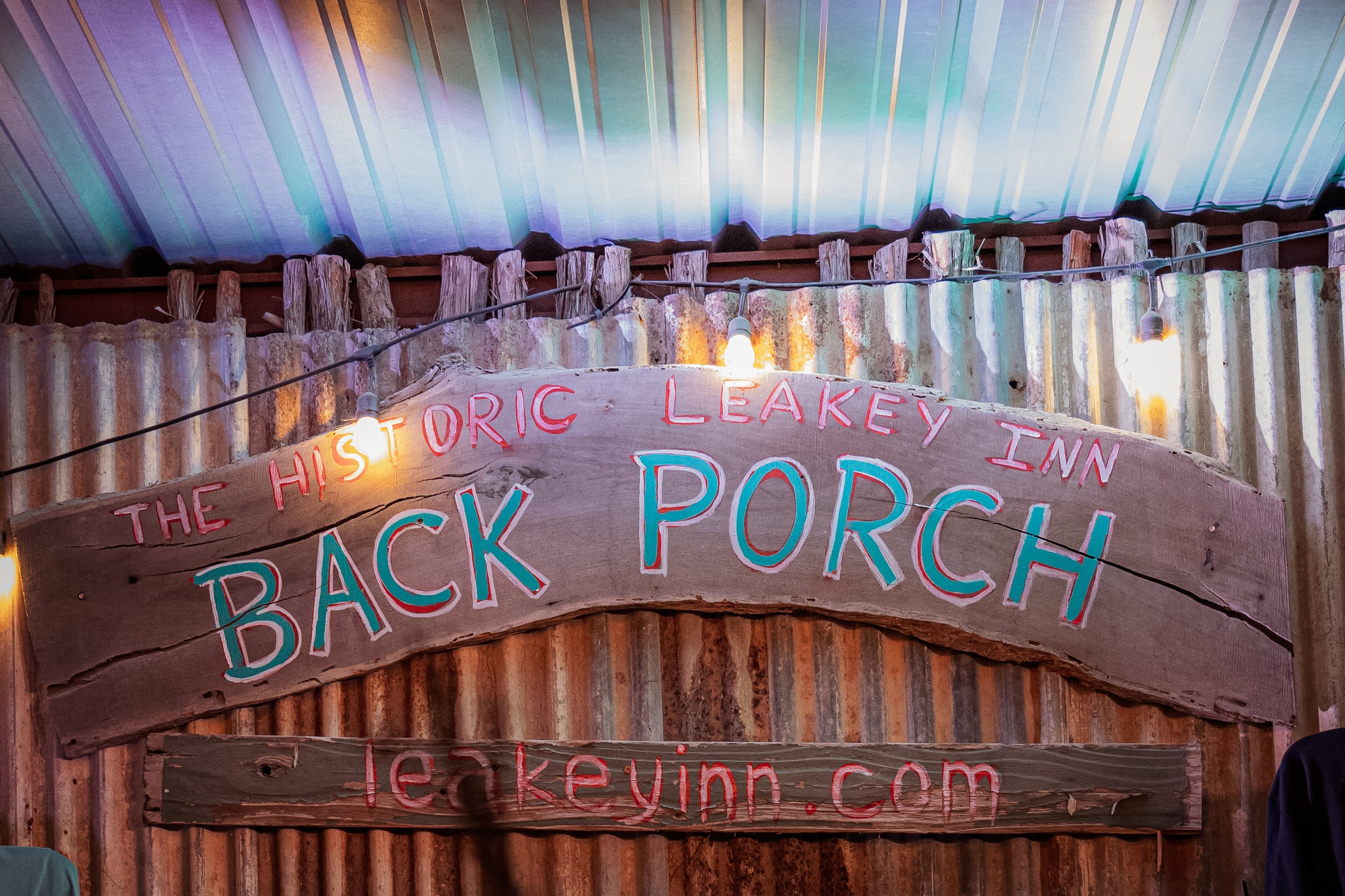 A rustic wooden sign with the words 'The Historic Leakey Inn Back Porch' and a website 'leakeyinn.com' painted in colorful letters, hanging above a corrugated metal wall with string lights and a colorful ceiling.