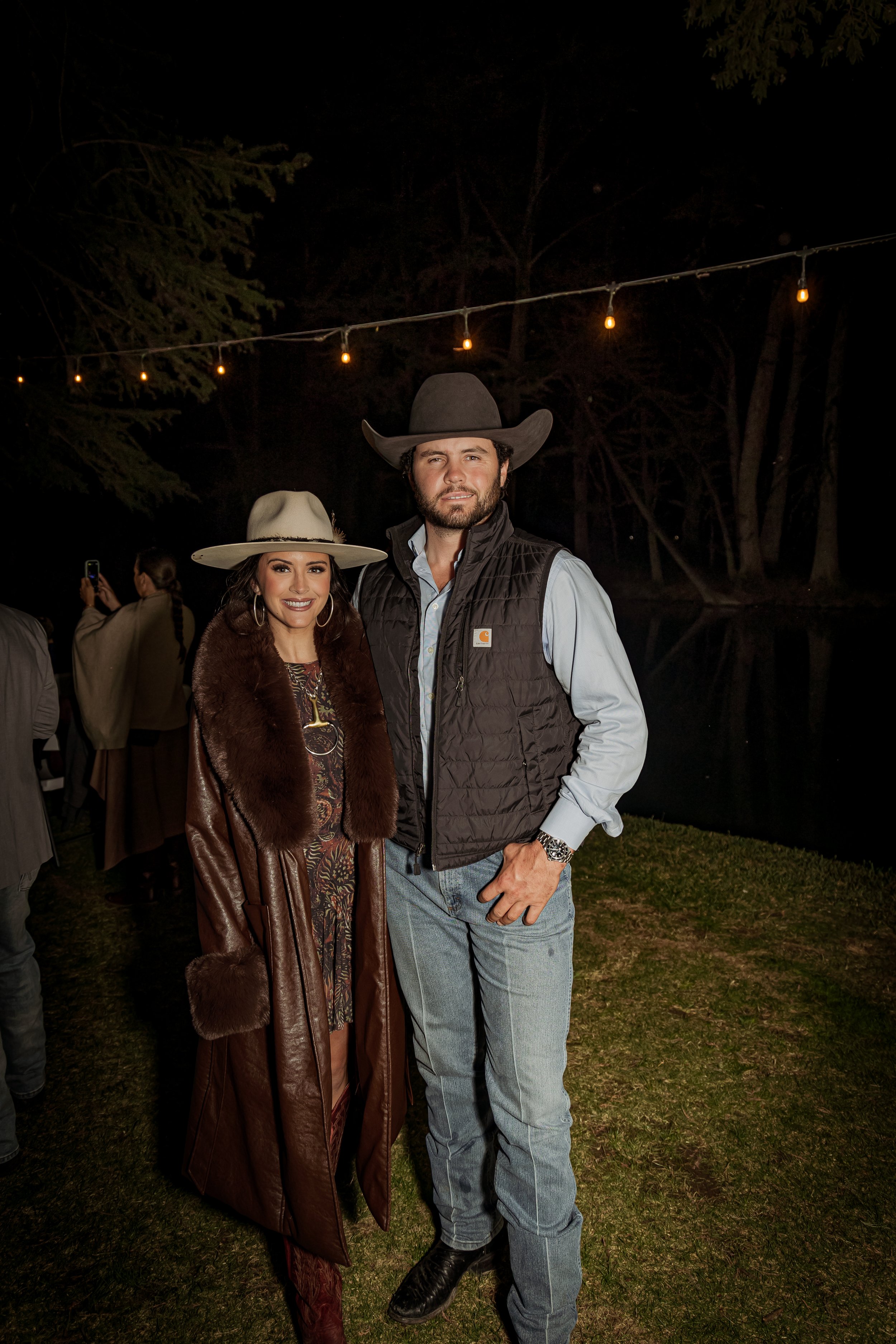 A man and woman dressed in western attire standing outdoors at night, illuminated by string lights overhead. The woman is wearing a wide-brimmed hat, a fur-collared leather coat, and patterned dress, while the man is wearing a cowboy hat, a light blu