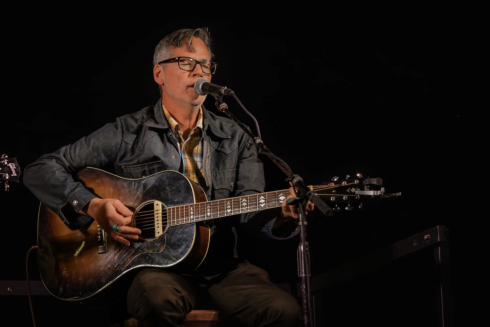 Man with glasses playing and singing into a microphone while seated and holding an acoustic guitar on stage with a black background.