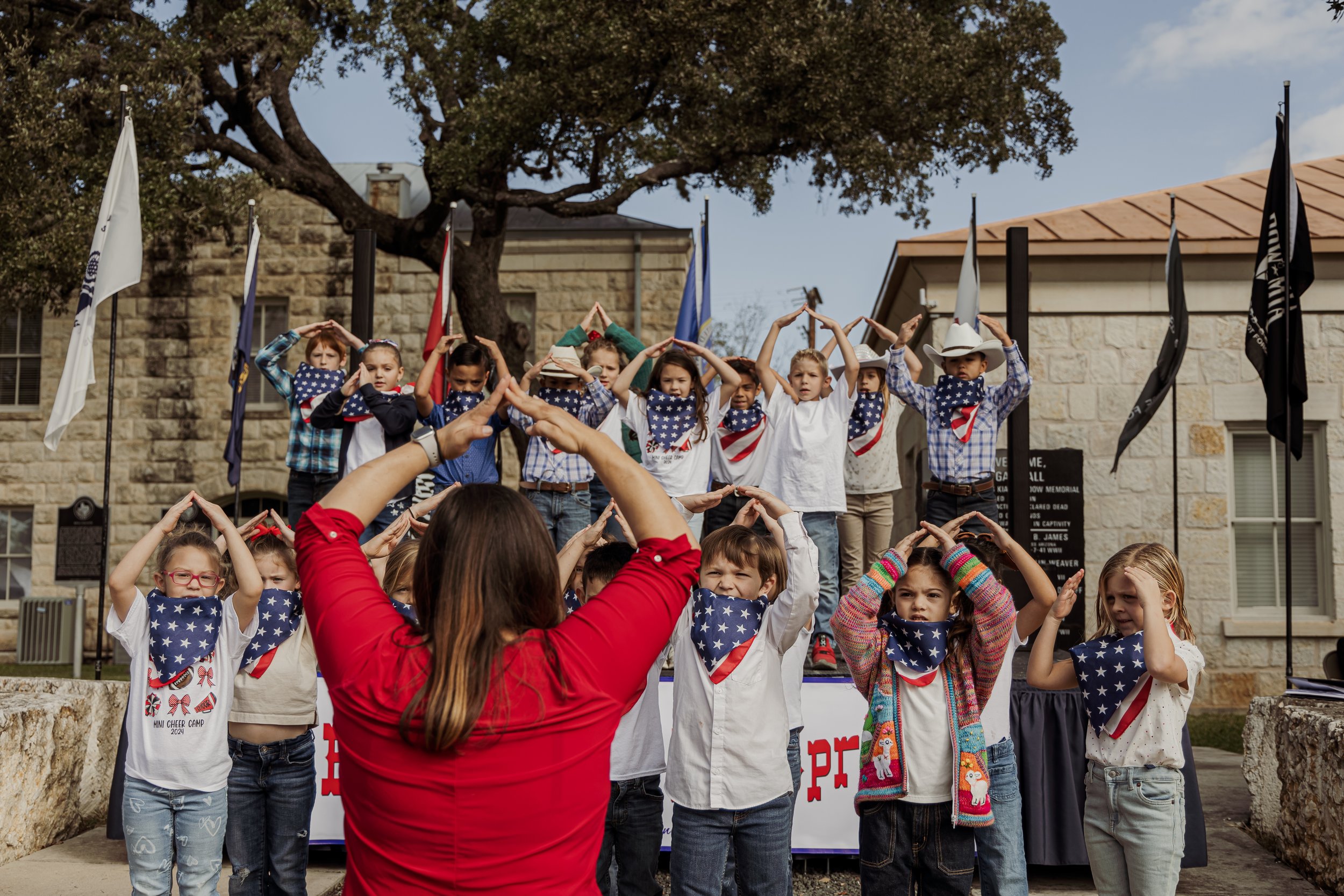 Children wearing American flag-themed bandanas participating in a Flag Day event, with some on an elevated platform and others on the ground, while a woman in a red shirt faces them with her back to the camera.