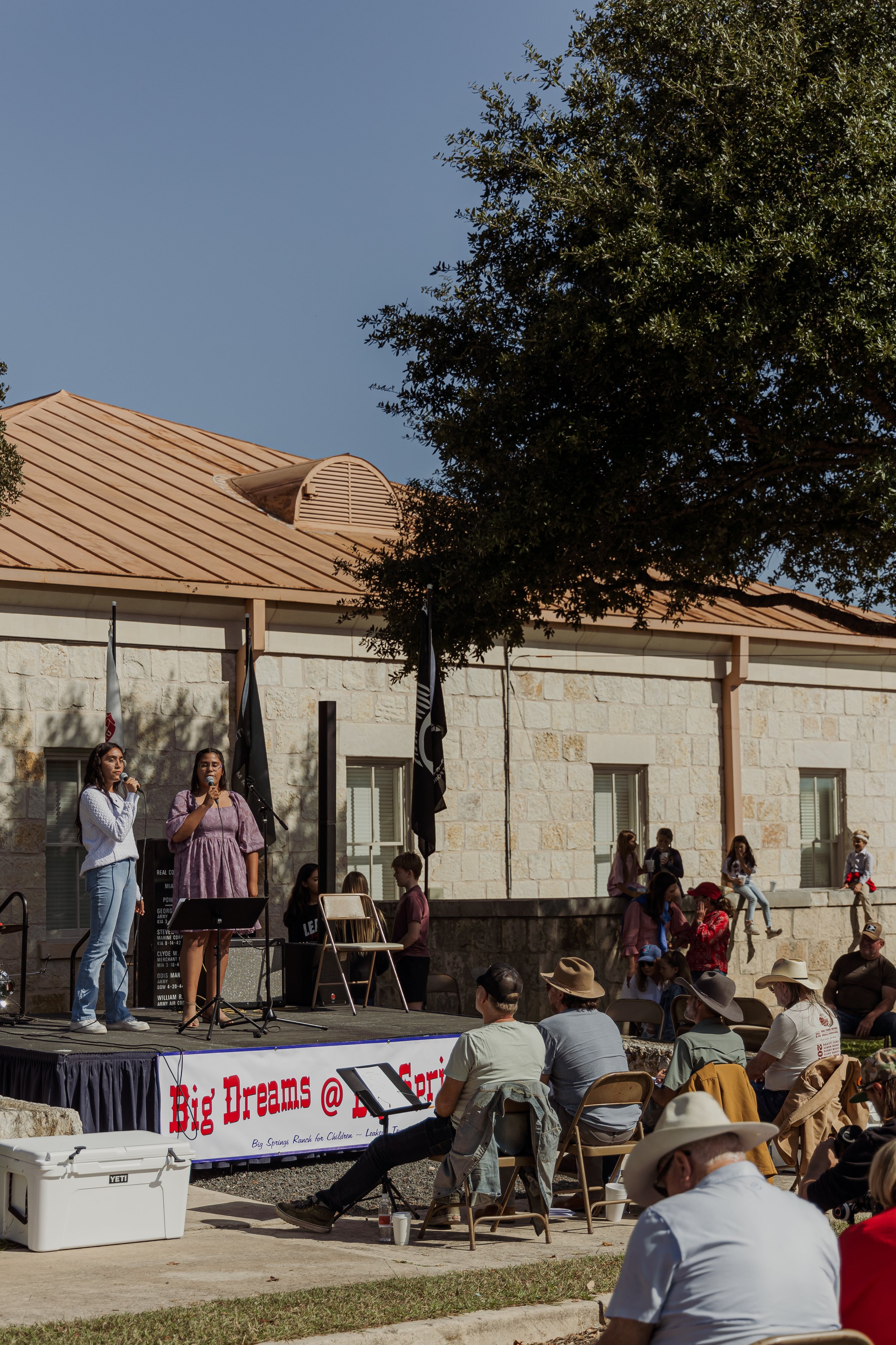 A crowd gathers in front of a small outdoor stage at a community event, with people sitting on chairs and standing, watching women singing into microphones, with a banner that reads "Big Dreams @ Spring," and a building with large windows and flags i