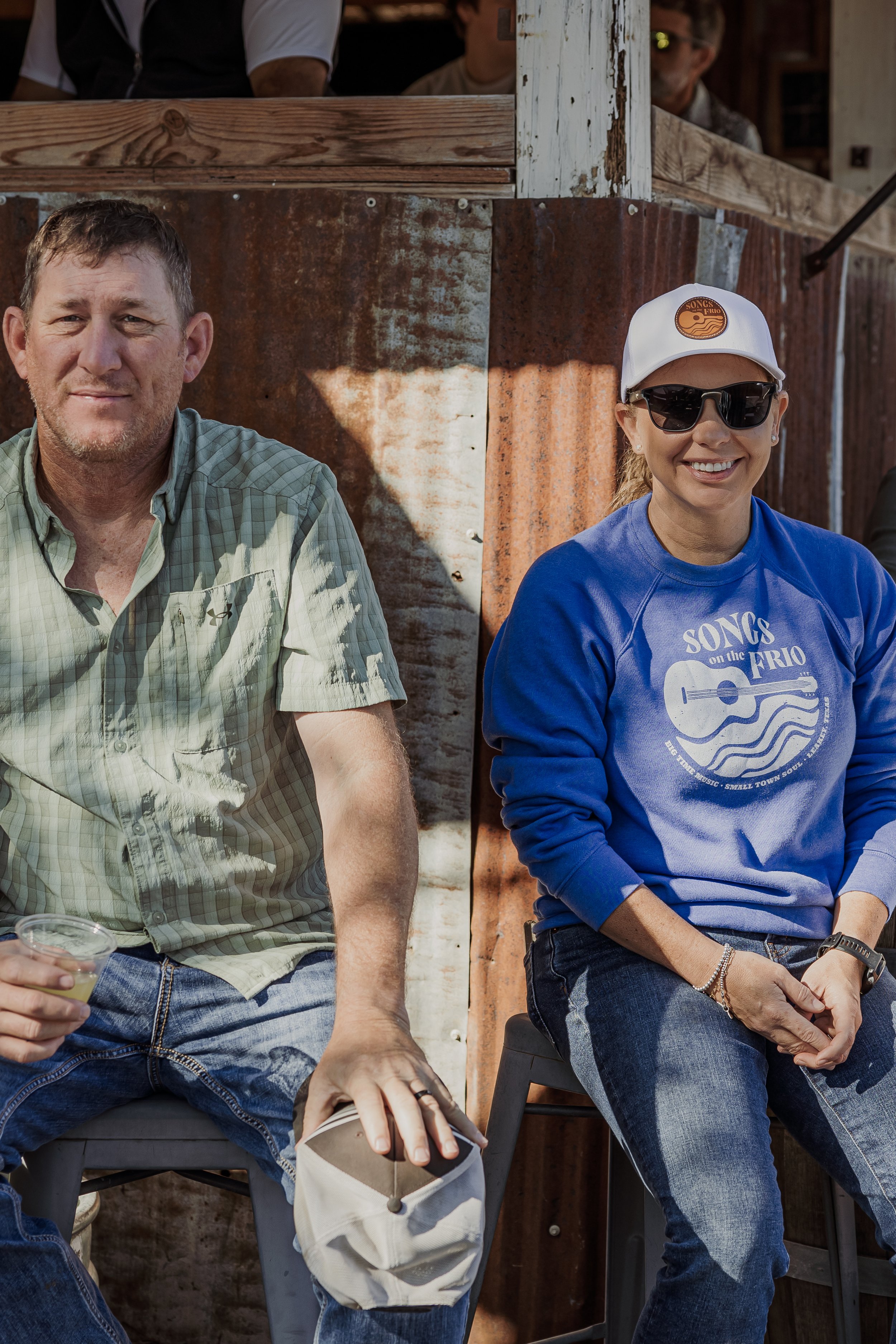 A man and woman sitting outdoors on chairs, smiling at the camera. The man is wearing a green checked shirt, holding a drink and a cap. The woman is wearing a blue sweatshirt, sunglasses, a white cap, and has jewelry on her wrists.