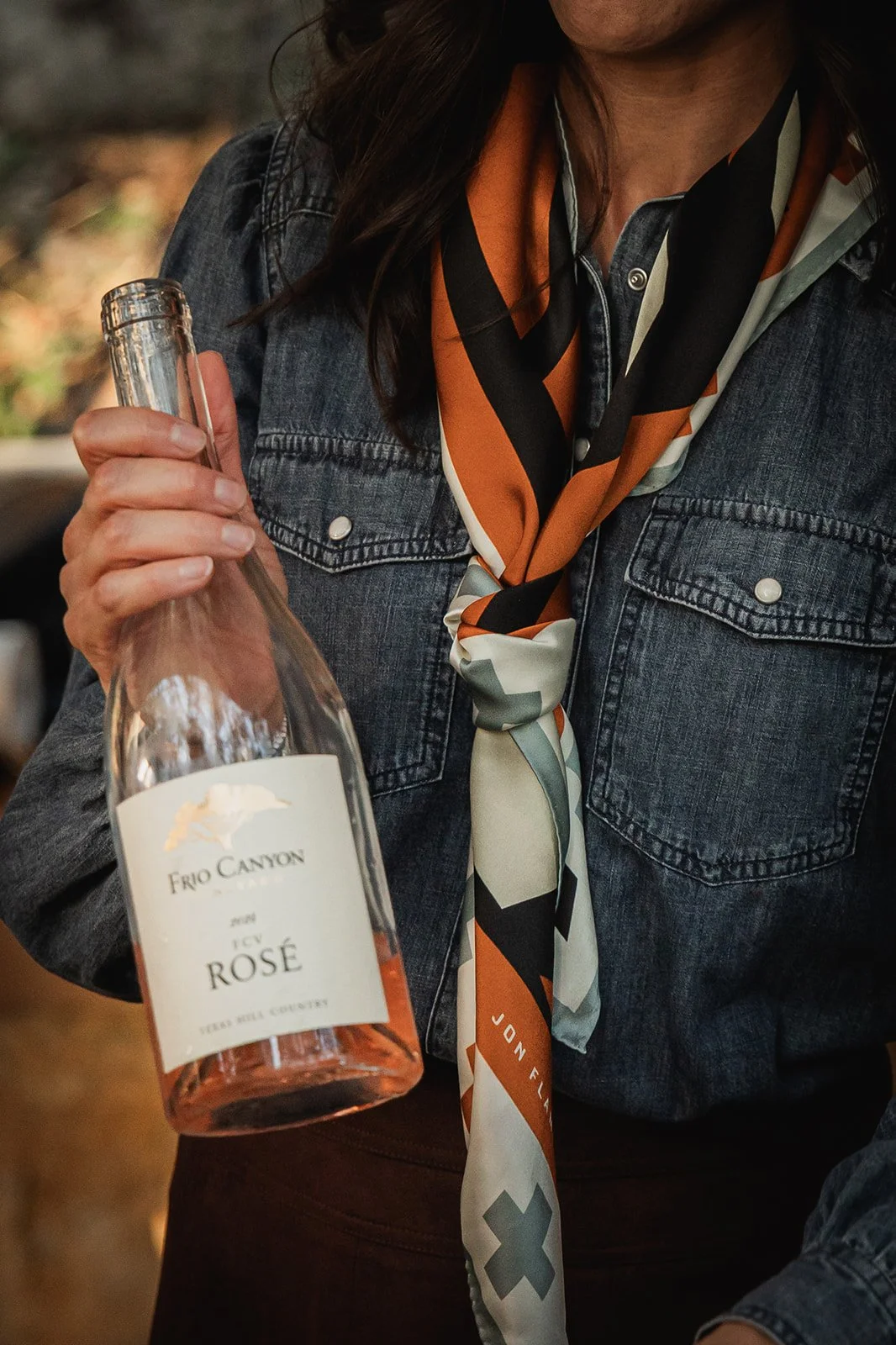 Person holding a bottle of Frio Canyon Rosé wine, wearing a denim jacket and a colorful scarf.