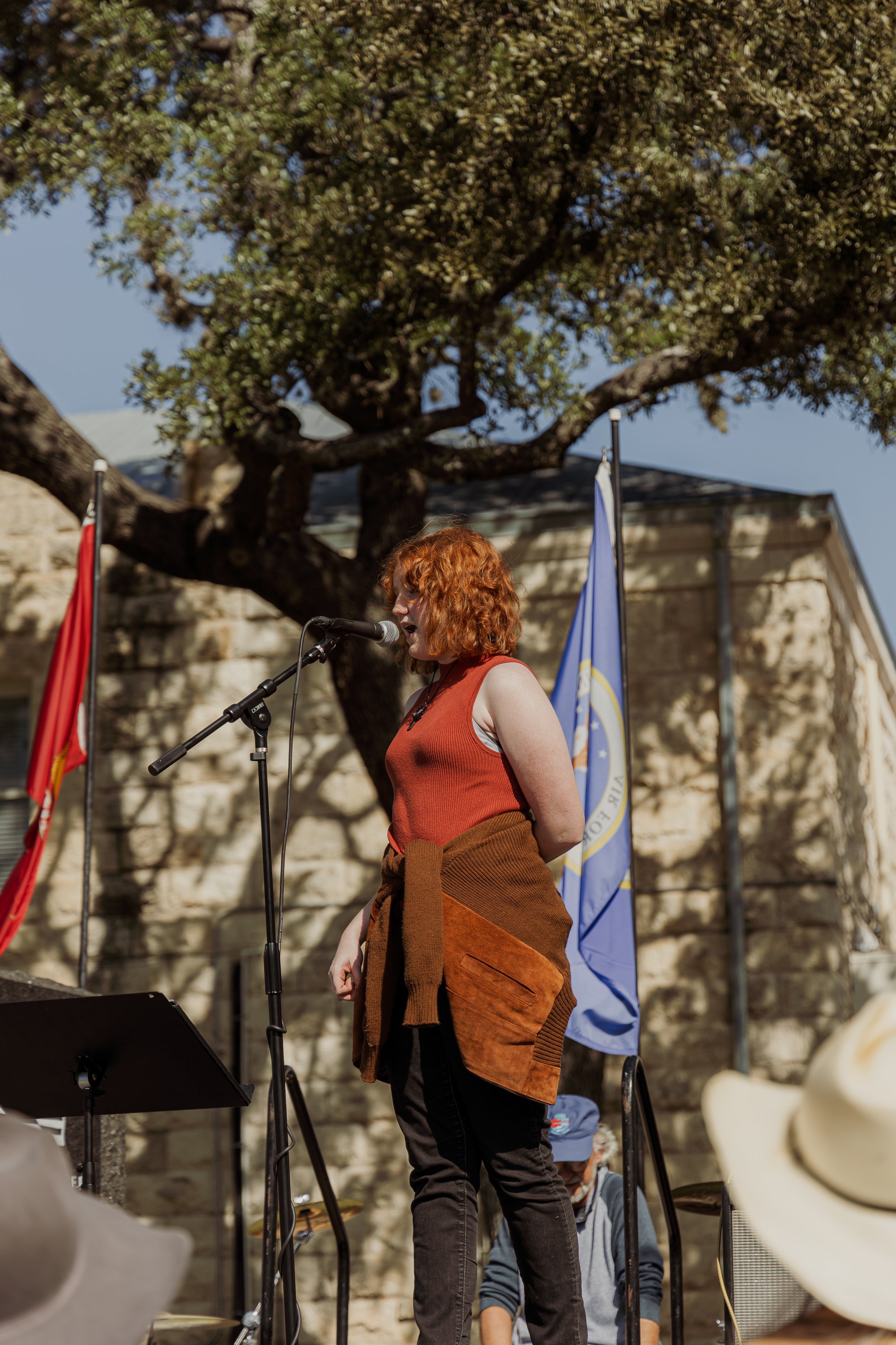 A woman with curly red hair in a sleeveless orange top speaking into a microphone at an outdoor event, with flags and a large tree in the background.