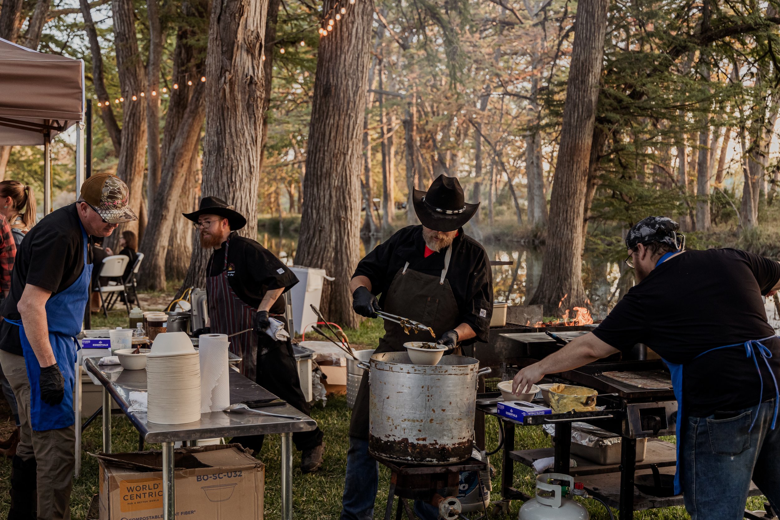 People cooking outdoors at a barbecue event in a park, with trees and string lights in the background.