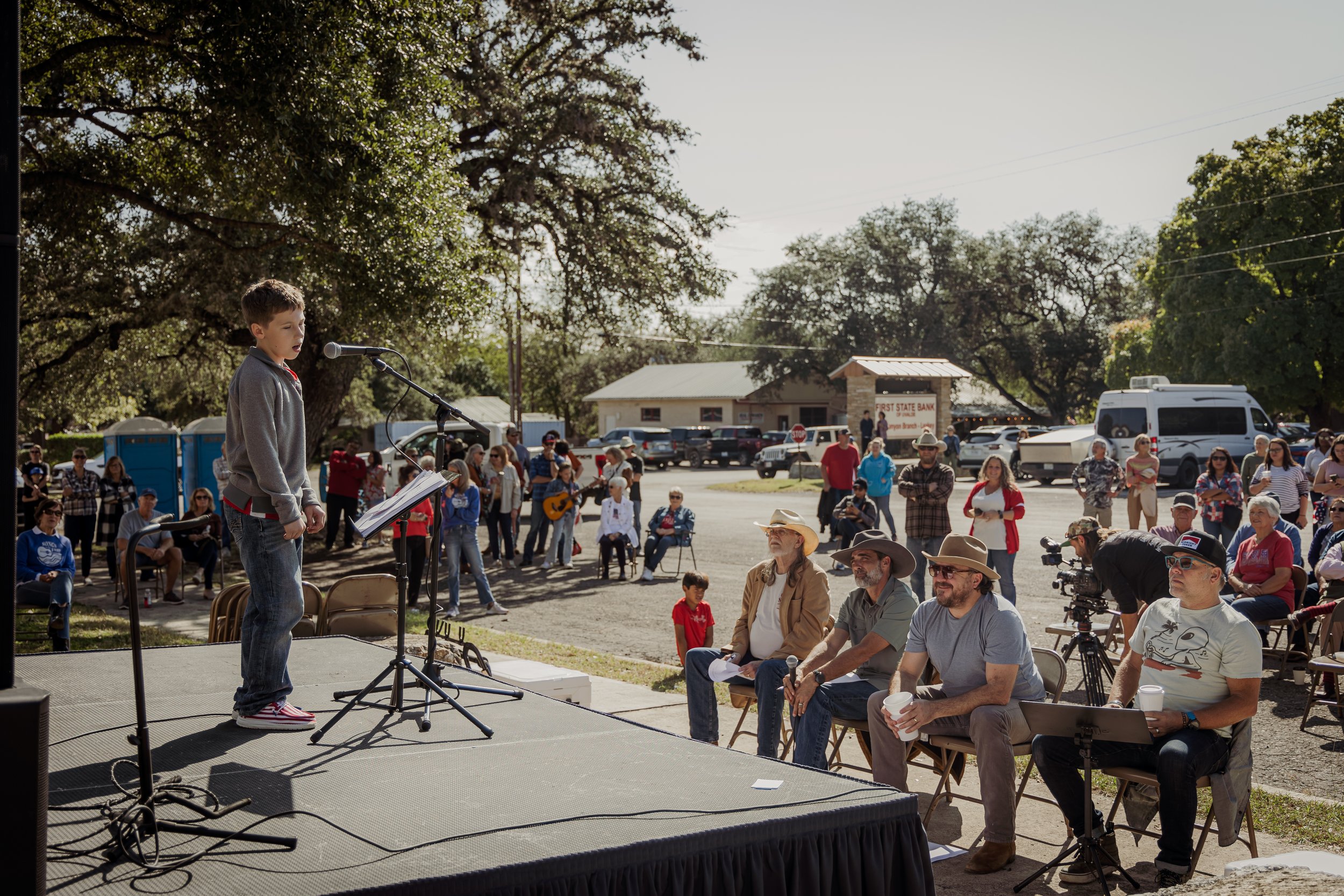 A young boy is speaking at a microphone on an outdoor stage during a community event, with an audience of adults and children sitting and standing around, some sitting in chairs and others standing behind. Trees and a building are visible in the back