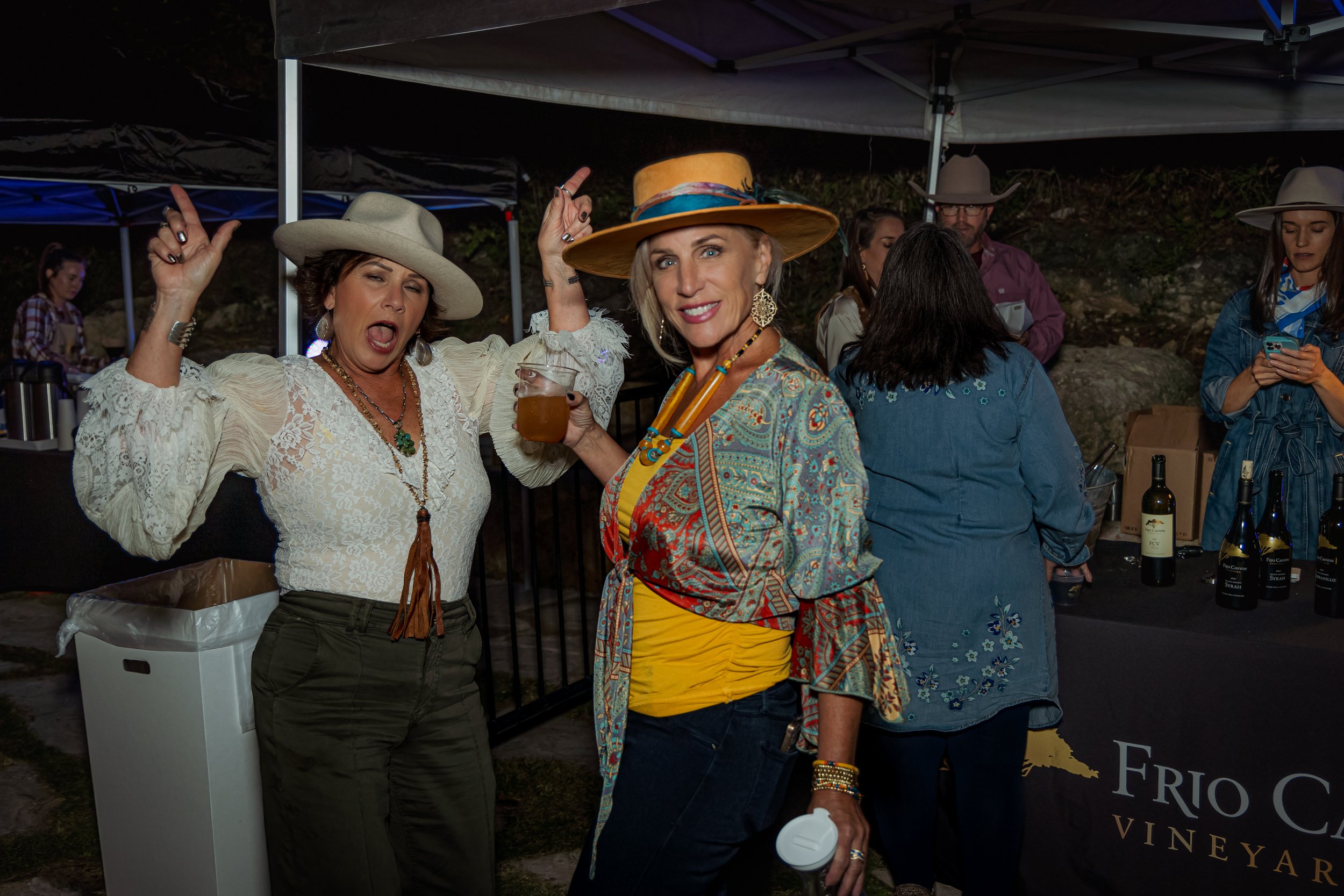 Two women dressed in western style at an outdoor vineyard event, one holding a drink and smiling, the other making a lively expression with her mouth open, both wearing hats and colorful clothing.