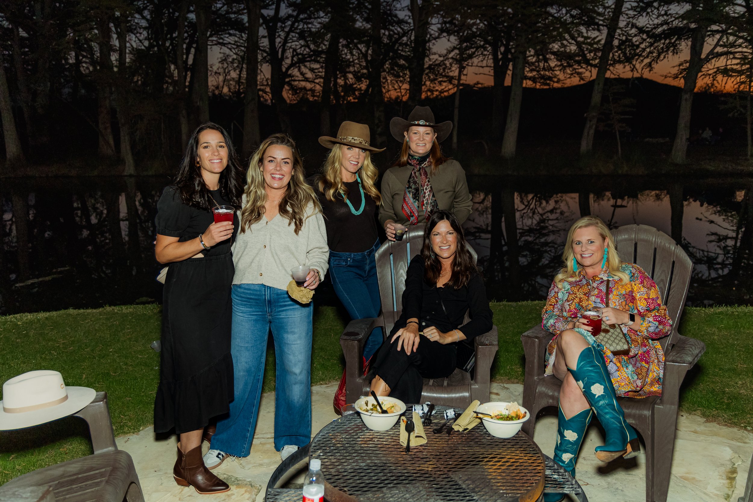 Group of six women at an outdoor evening gathering, with trees and a lake in the background. Some women are standing, others are seated, holding drinks, smiling. They are dressed in casual and western-inspired clothing.