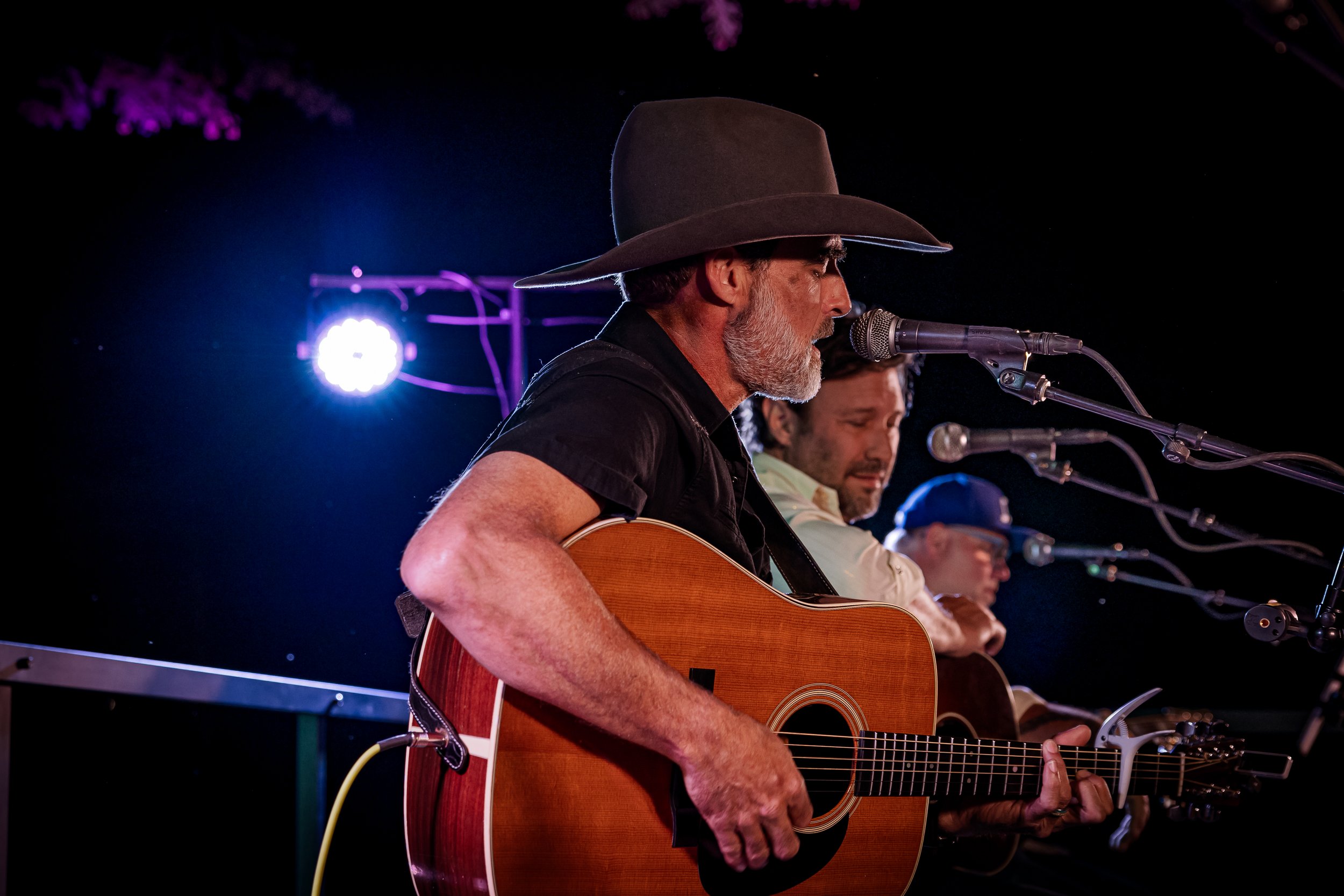 A man with a gray beard wearing a wide-brimmed hat and black shirt playing an acoustic guitar at a live performance, with two other musicians also singing into microphones. The stage is lit with purple and white lights.