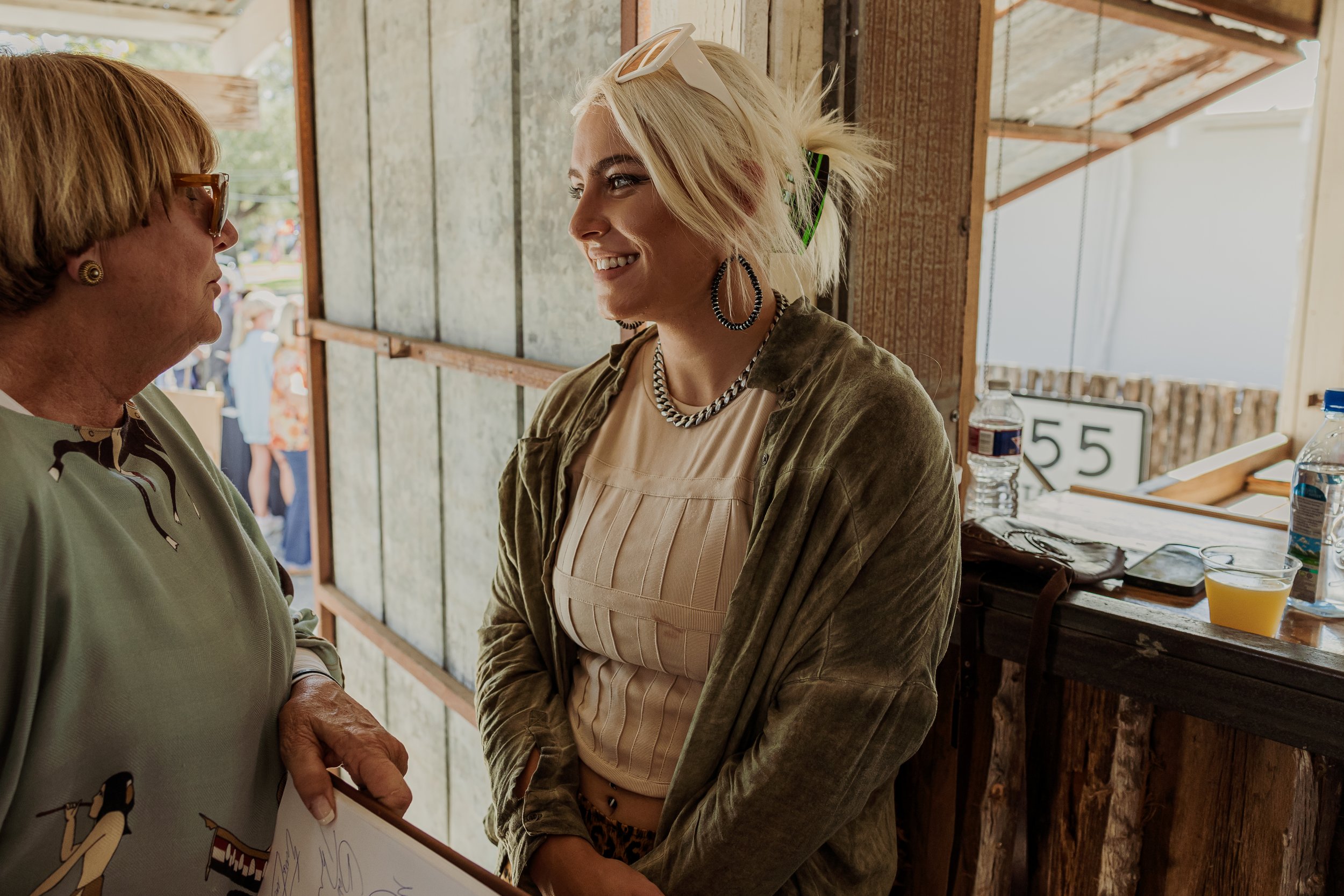 A woman with blonde hair, ice sunglasses, and jewelry smiling and talking to an older woman with short light brown hair and glasses inside a rustic wooden structure.