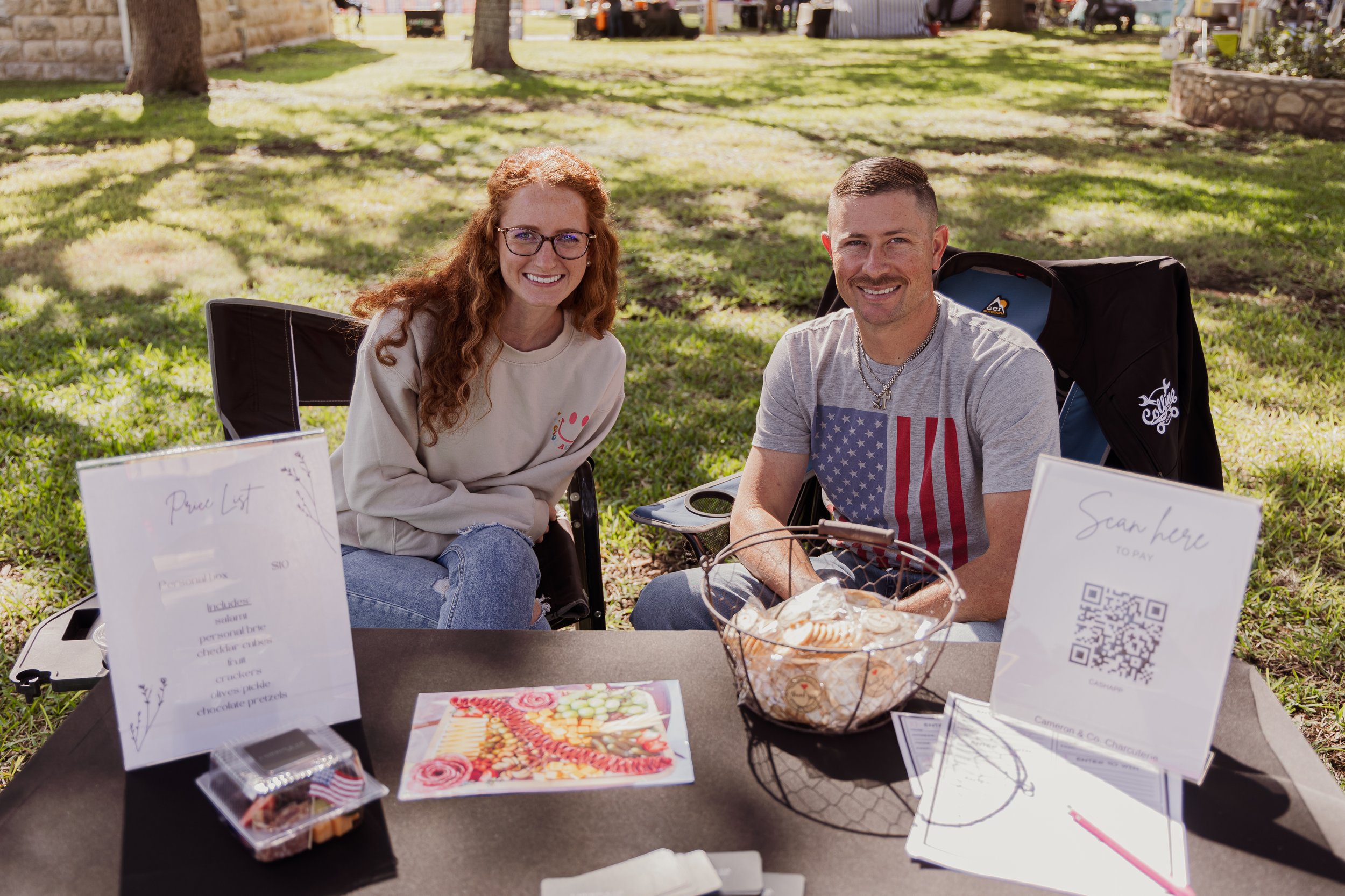 A man and a woman sitting at a table outdoors, smiling at the camera, with signs, a bowl of cookies, and artwork on the table, during a daytime event in a park with trees and green grass.