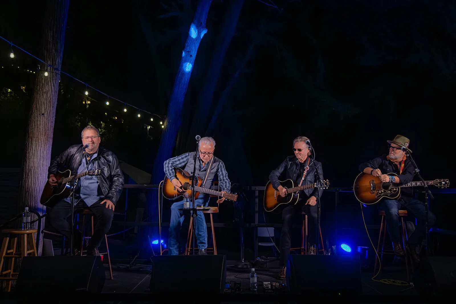 Four male musicians sitting on stage outdoors at night, each playing an acoustic guitar. They are illuminated by stage lights, with trees and string lights visible in the background.