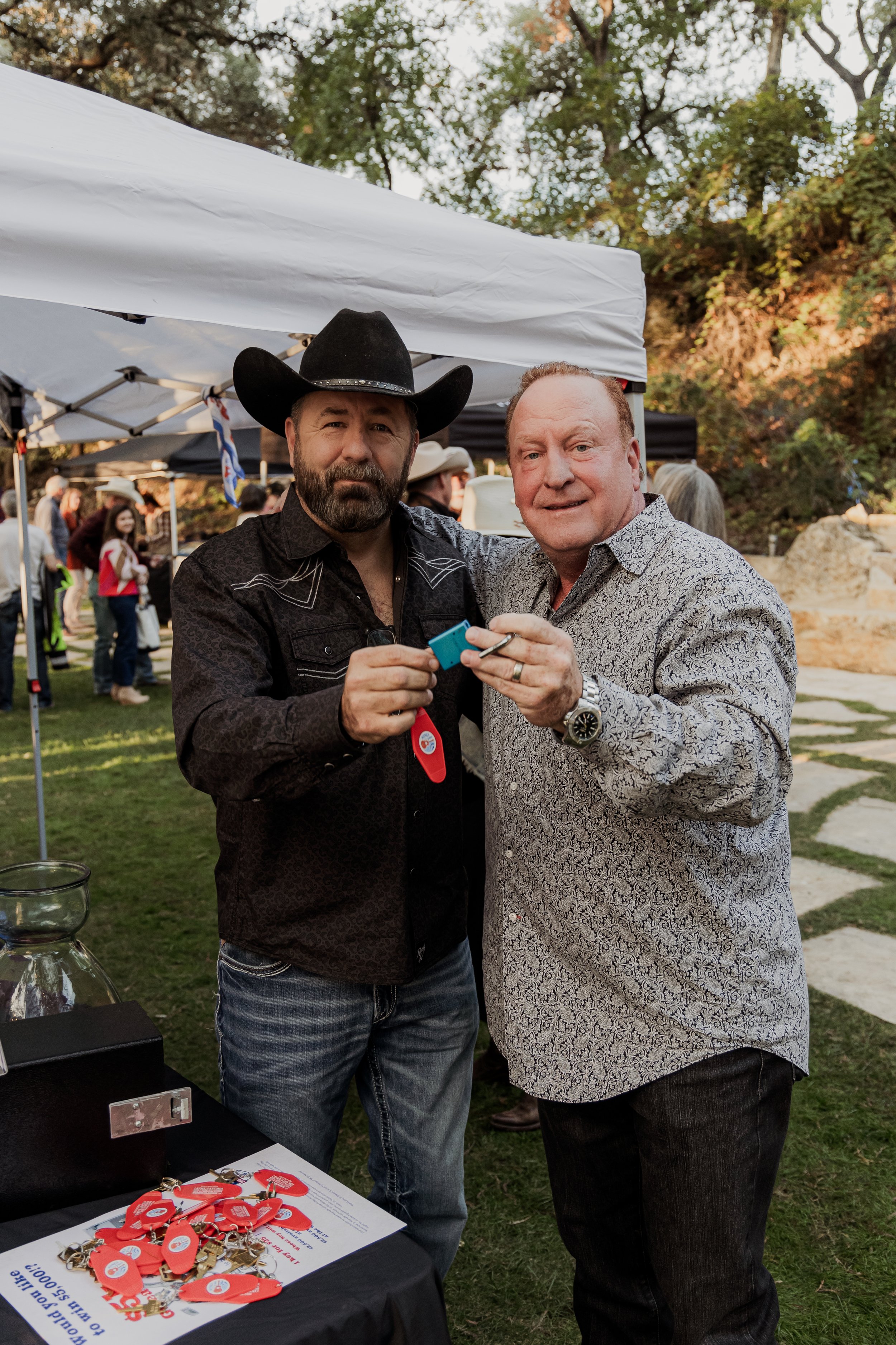 Two men, one wearing a cowboy hat and the other in a patterned shirt, stand together outdoors at an event, holding small blue objects, with a table of red and gold keychains in front of them and other people and tents in the background.