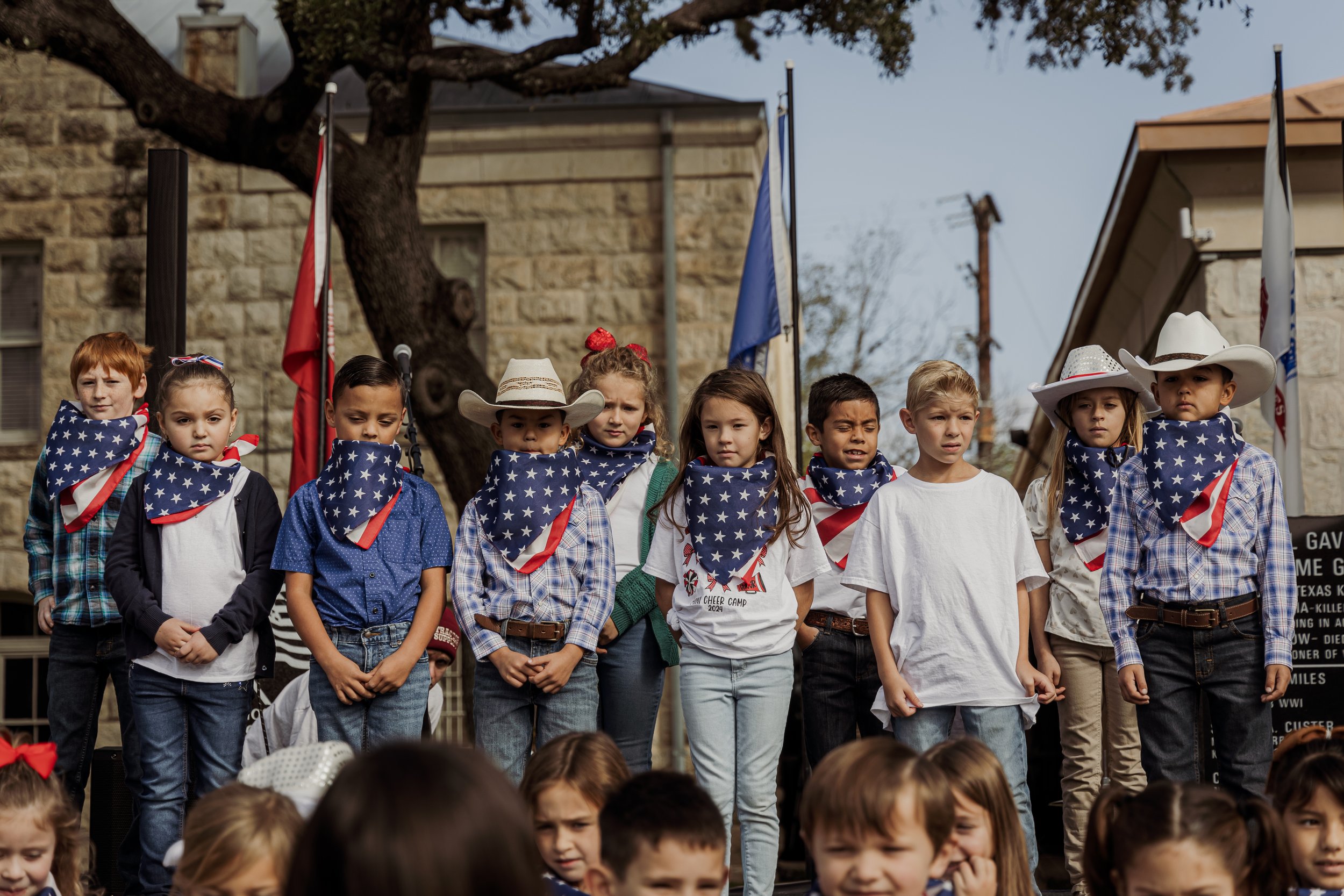 Children dressed in Western-style clothing, wearing American flag bandanas, gathered on a stage for a patriotic event, with flags and a large tree in the background.