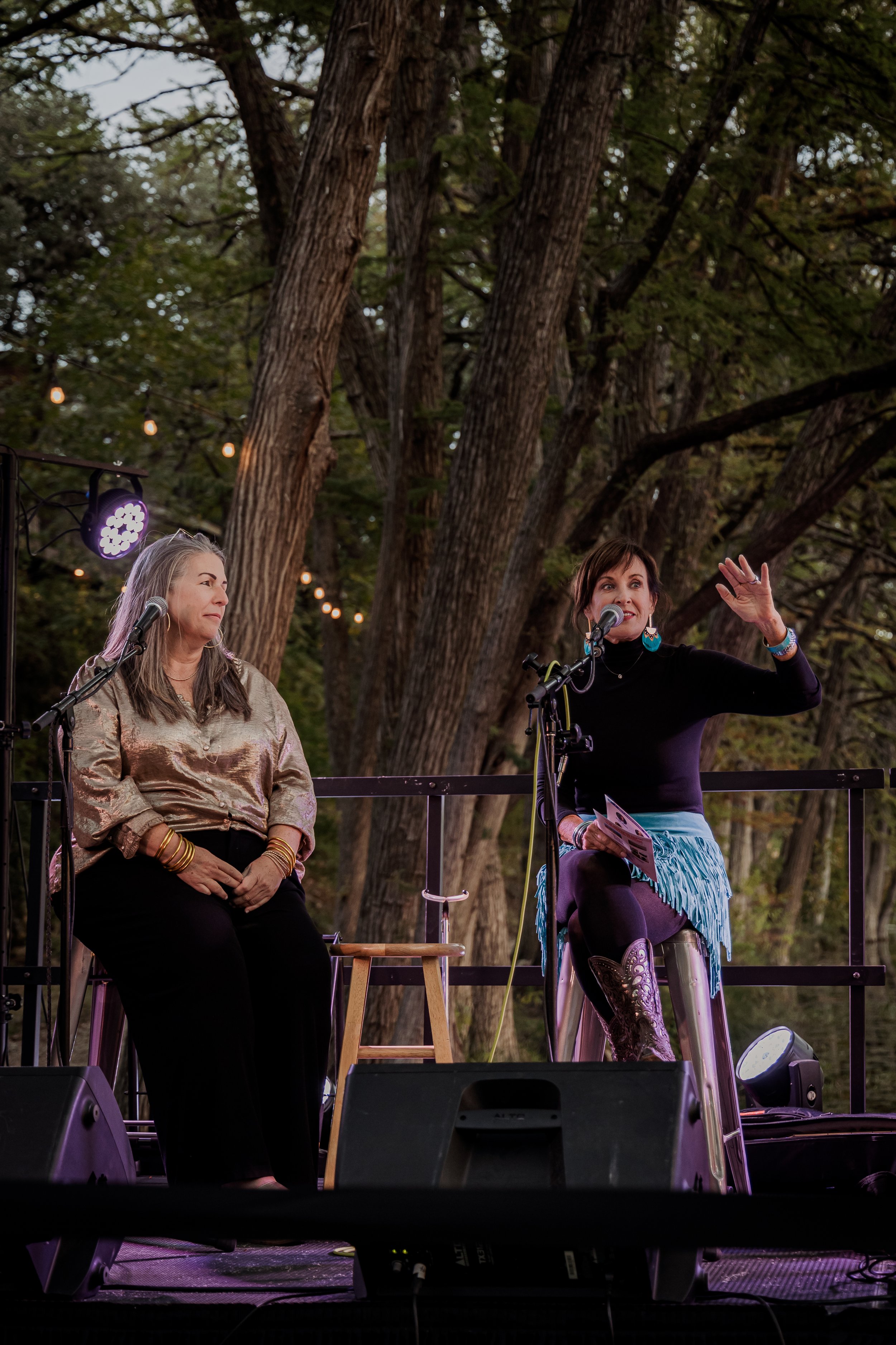 Two women on an outdoor stage between large trees, one speaking into a microphone and the other sitting beside her, with string lights above.
