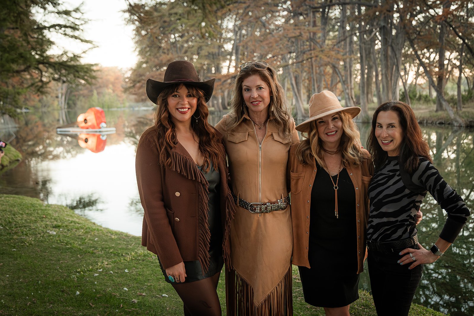 Four women standing together near a lake with trees and a boat in the background, dressed in casual autumn clothing and hats, smiling at the camera.