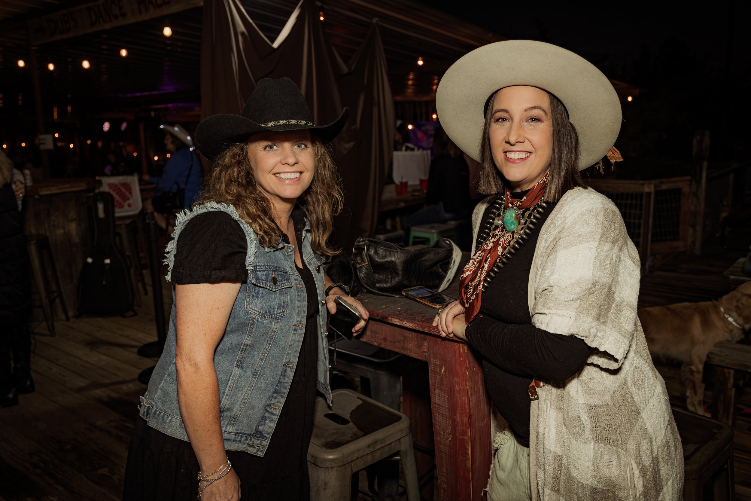 Two women wearing cowboy hats and Western attire stand inside a rustic venue, smiling at the camera.