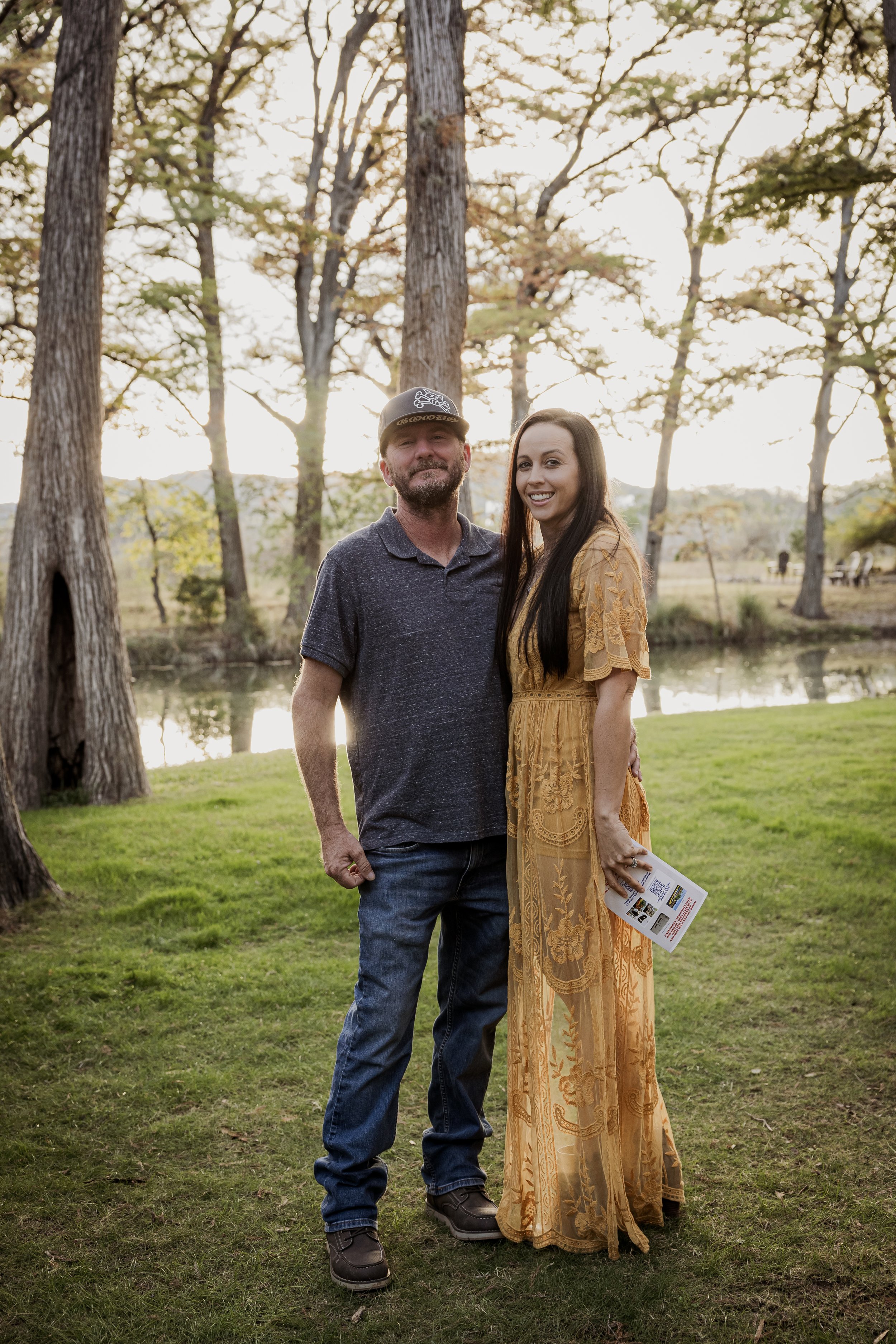 A man and woman standing together outdoors near a pond, smiling at the camera during sunset, with trees and a grassy area in the background.