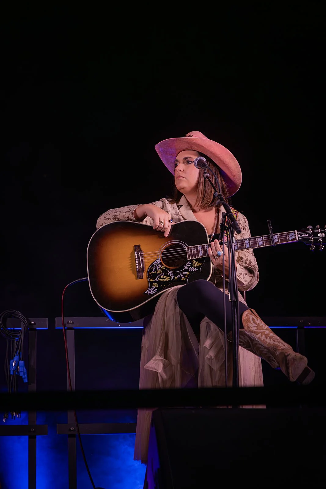 A woman wearing a pink cowgirl hat and vintage clothing, sitting on a stool, playing an acoustic guitar on stage in front of a black background.