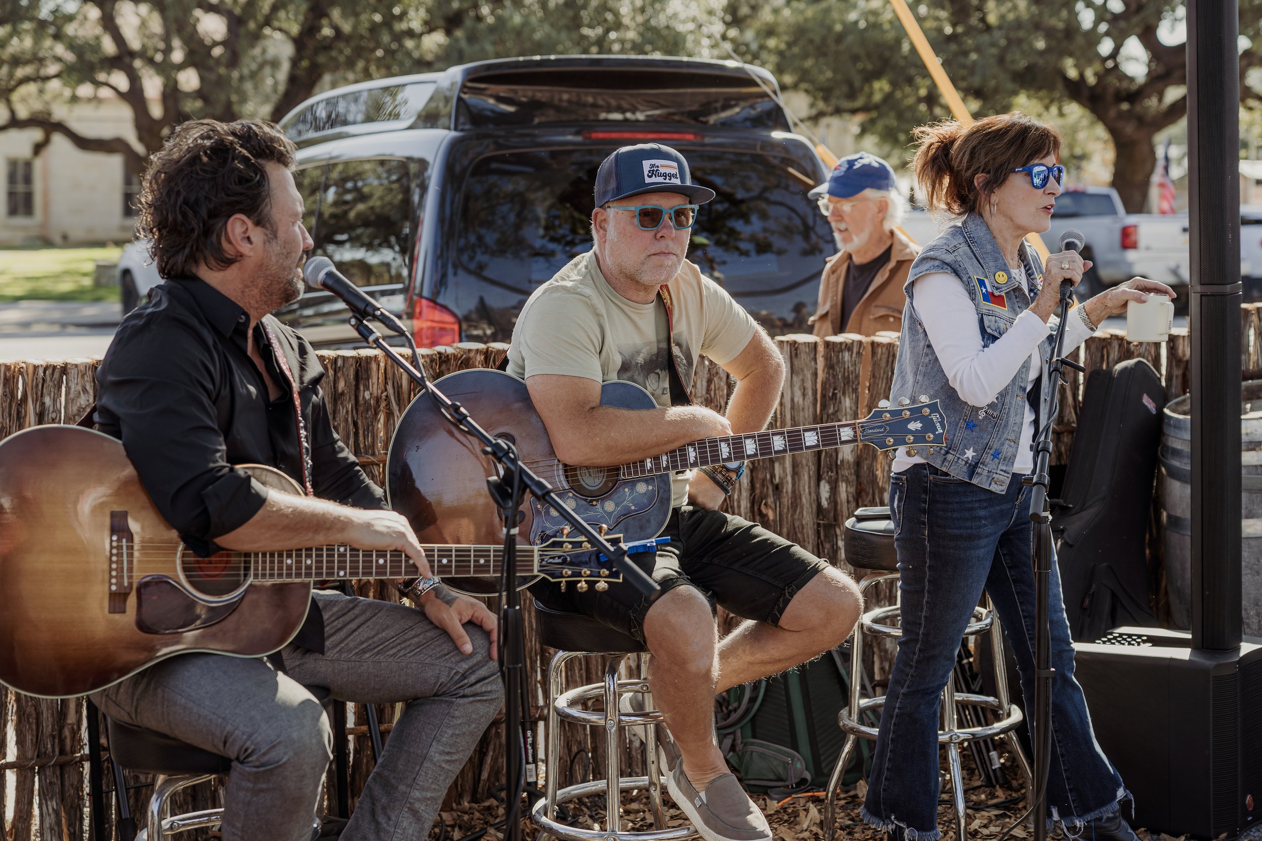 A band performing outdoors with two men playing acoustic guitars and a woman singing into a microphone, all sitting on stools in front of a wooden barrier, with cars and trees in the background.