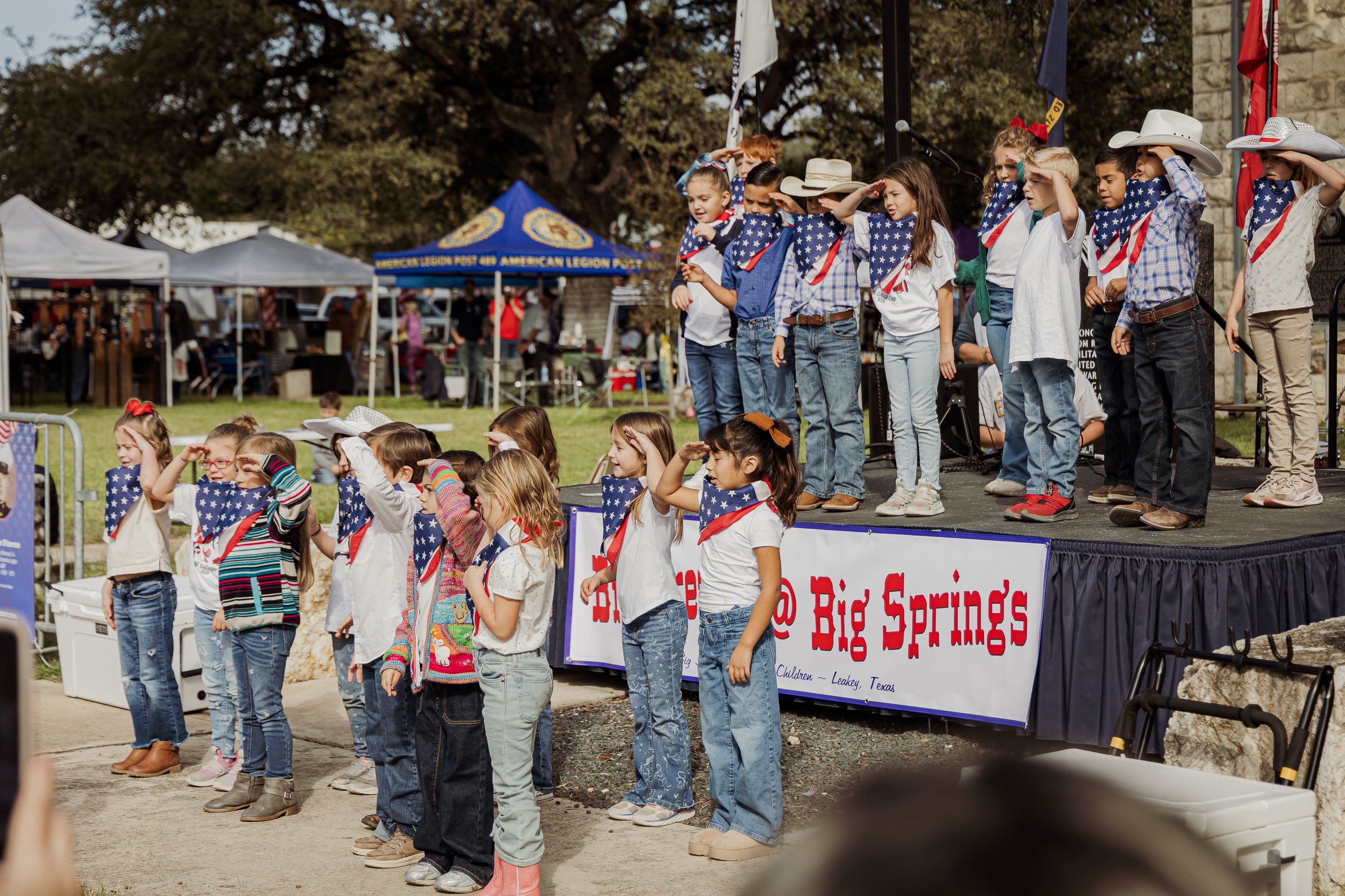 Children saluting and singing on a stage and in front of it, dressed with American flag-themed bandanas and clothes, during a patriotic event at Big Springs.
