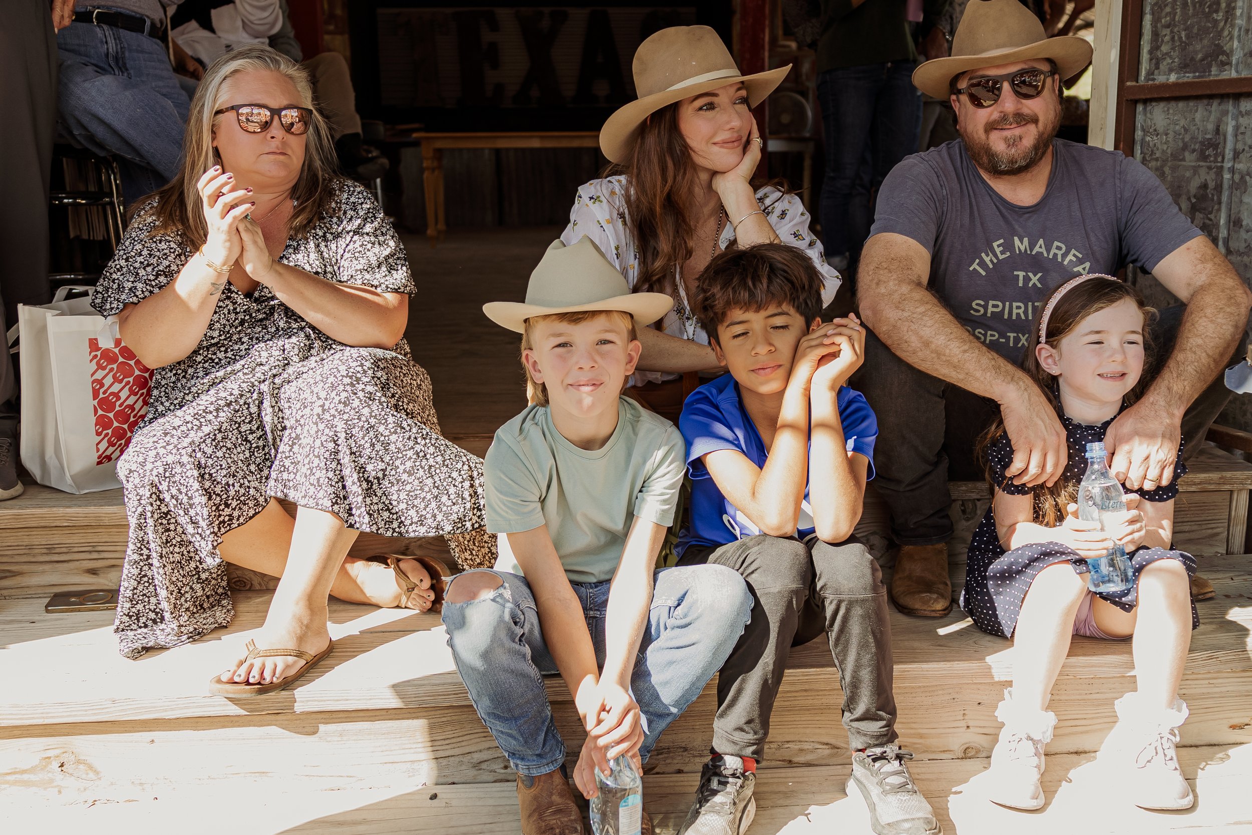 Group of people sitting on wooden stairs outdoors, including two women, two men, and three children, some wearing hats, enjoying a sunny day.