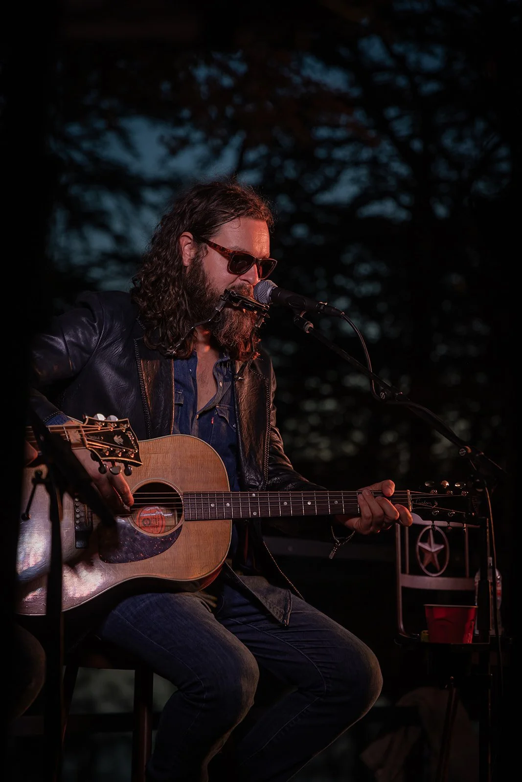 A man with long hair and a beard wearing sunglasses, a leather jacket, and a blue shirt, playing an acoustic guitar and singing into a microphone during an outdoor performance at dusk.