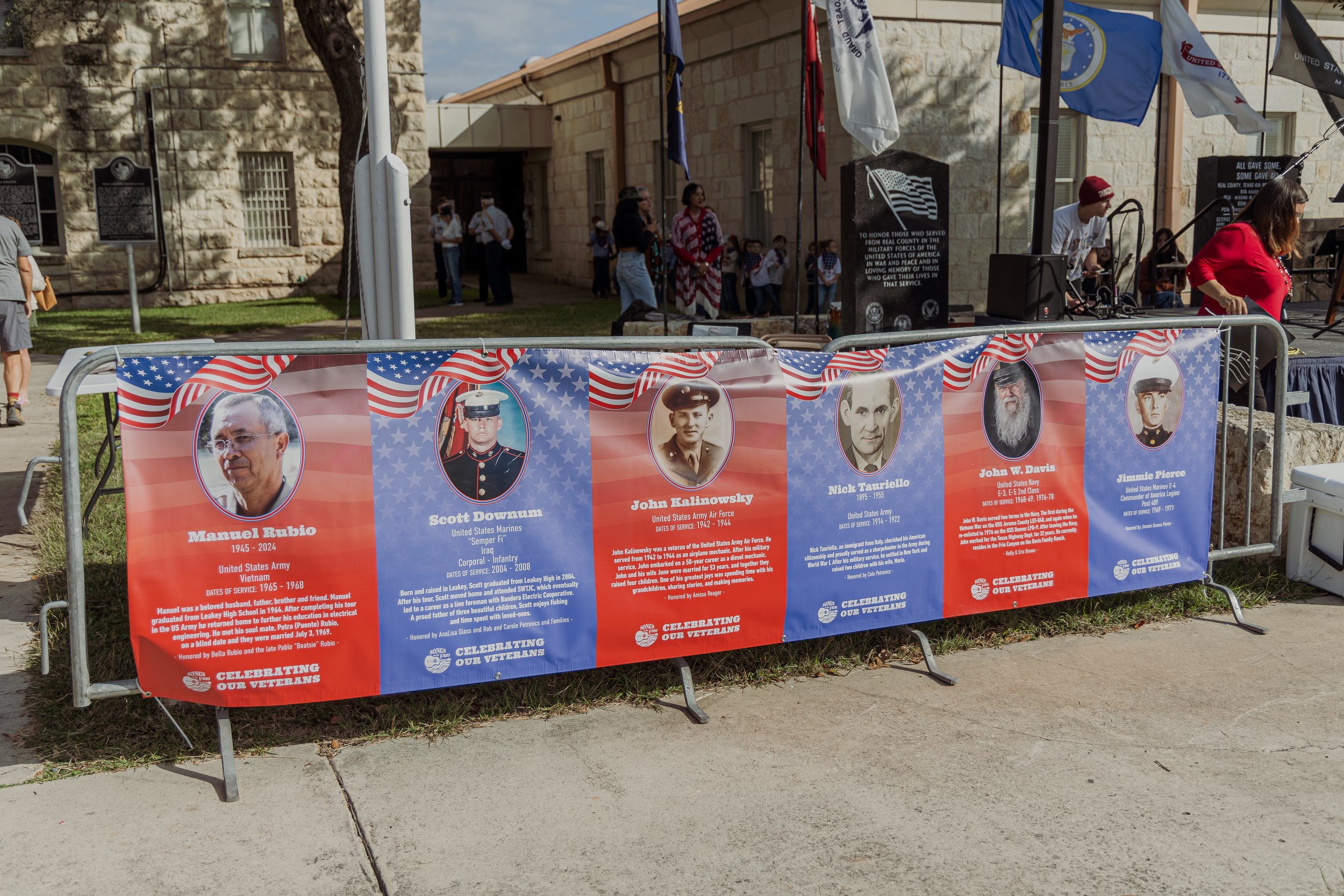 A patriotic memorial display honoring fallen military veterans, featuring large posters of five men with their names, photos, and service details, behind a metal barricade decorated with American flag graphics, set outdoors at a public event.