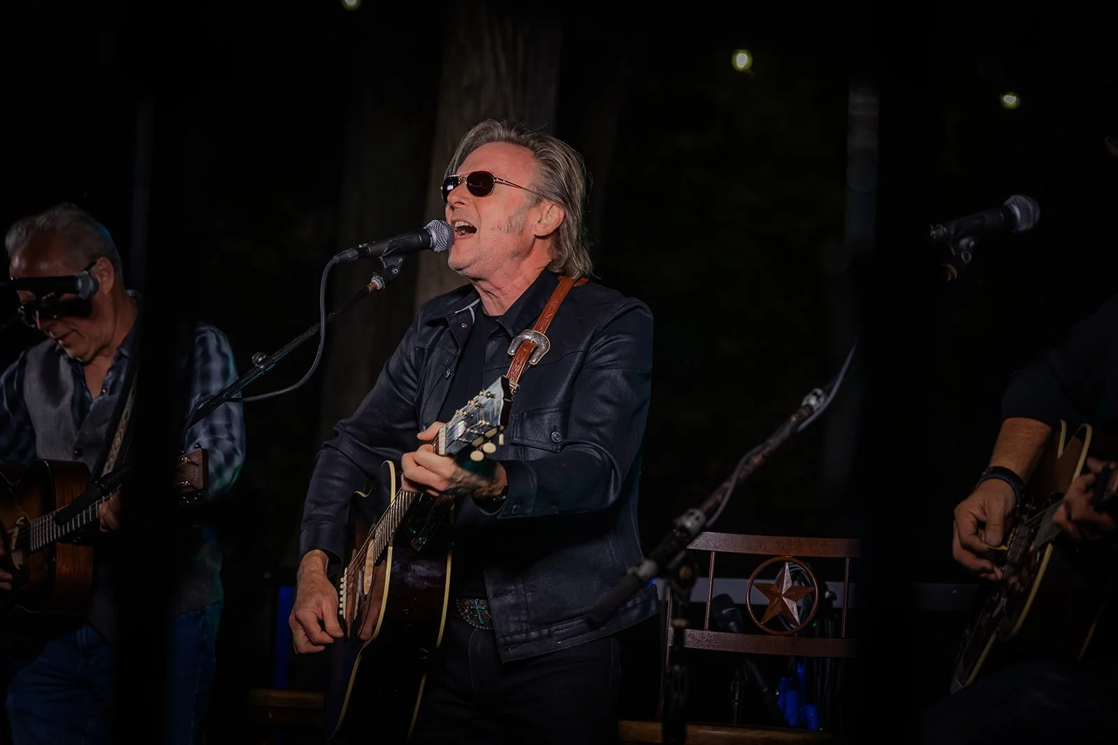 Musician with sunglasses singing and playing guitar during an outdoor performance at night, with other guitarists visible on stage.