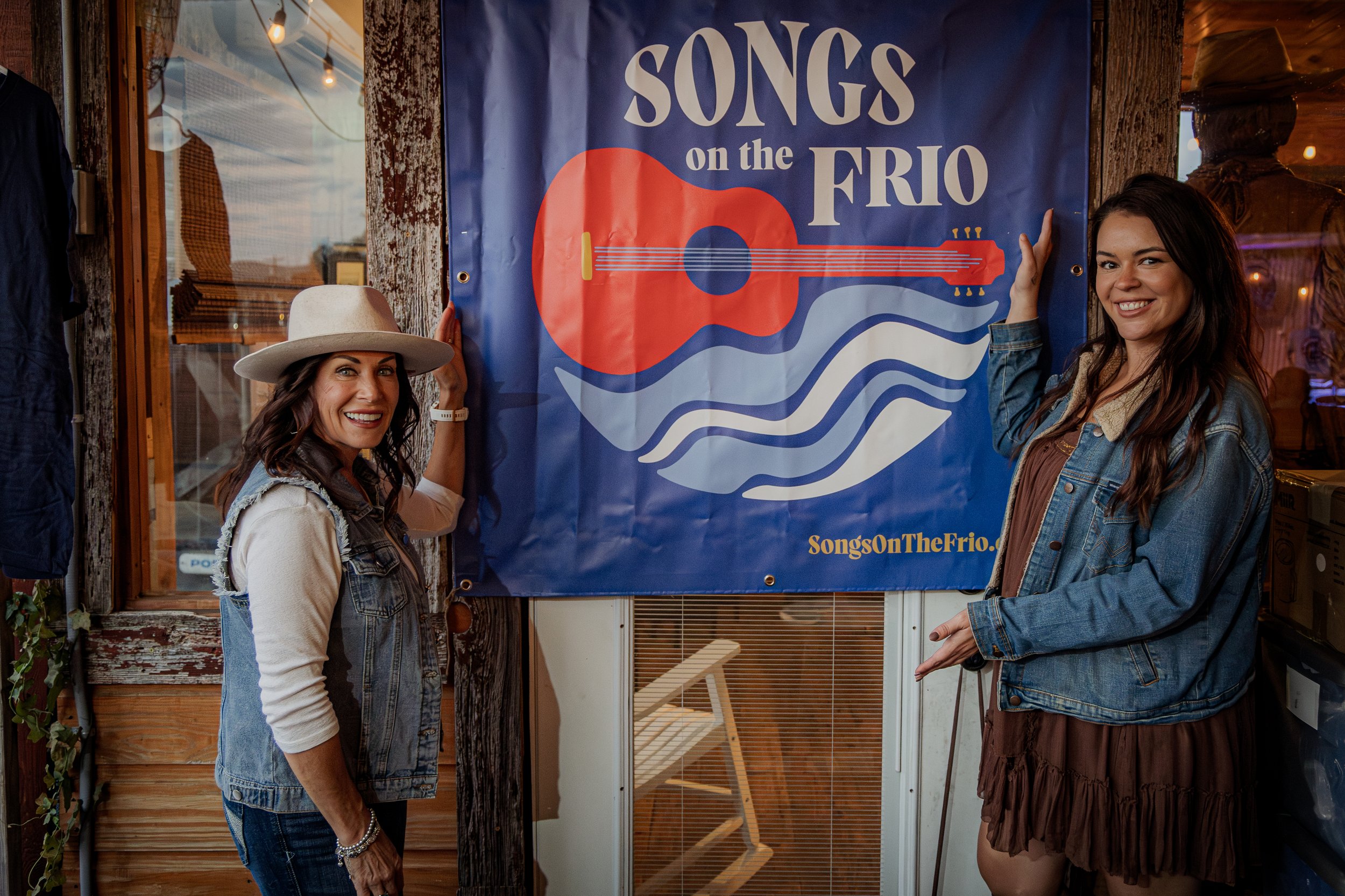 Two women smiling and standing next to a large blue banner that says 'Songs on the Frio' with a guitar illustration. One woman is wearing a wide-brimmed hat and denim vest, and the other is wearing a denim jacket and a brown dress. They are indoors,