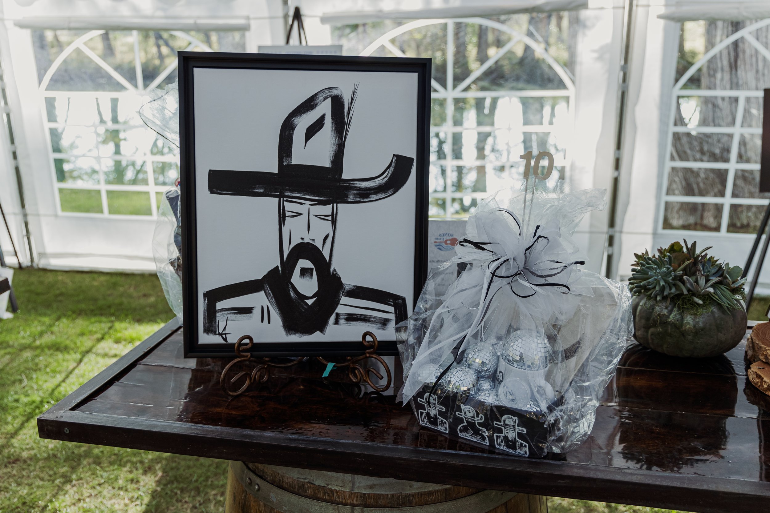 A black and white portrait of a man with a wide-brimmed hat, beard, and mustache, displayed on an easel. Next to the portrait is a clear plastic bag with small disco ball ornaments and a white ribbon with a black bow, marked with a gold