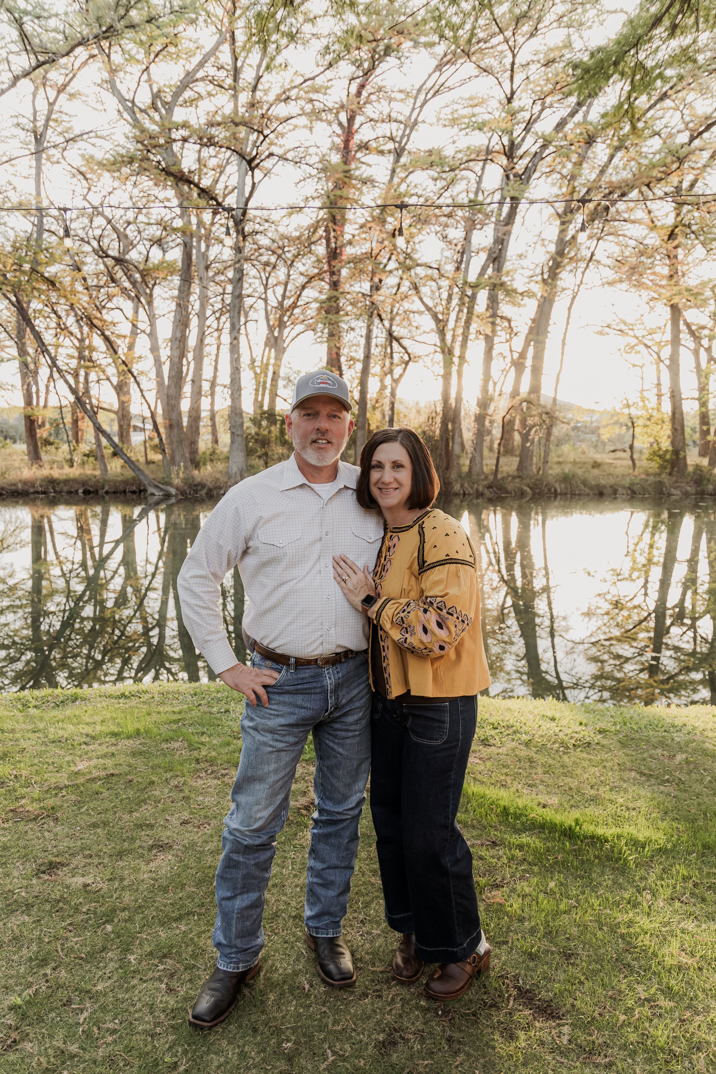 A middle-aged couple stands close together by a river, smiling at the camera during sunset, with trees in the background.