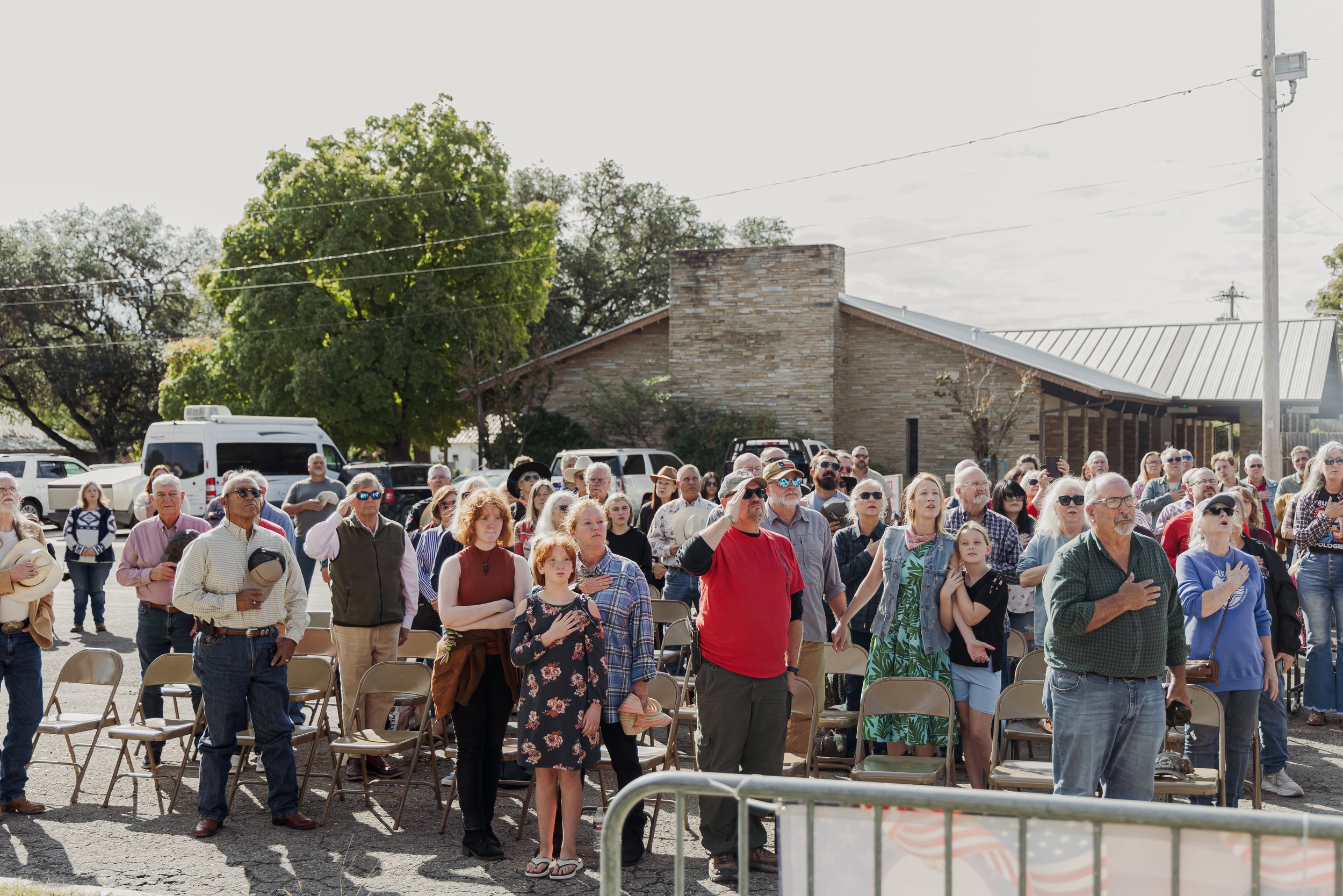 A large group of people gathered outdoors on a sunny day, standing and some seated in front of a building, with some holding their right hands over their hearts. Many appear to be participating in a patriotic or memorial event.