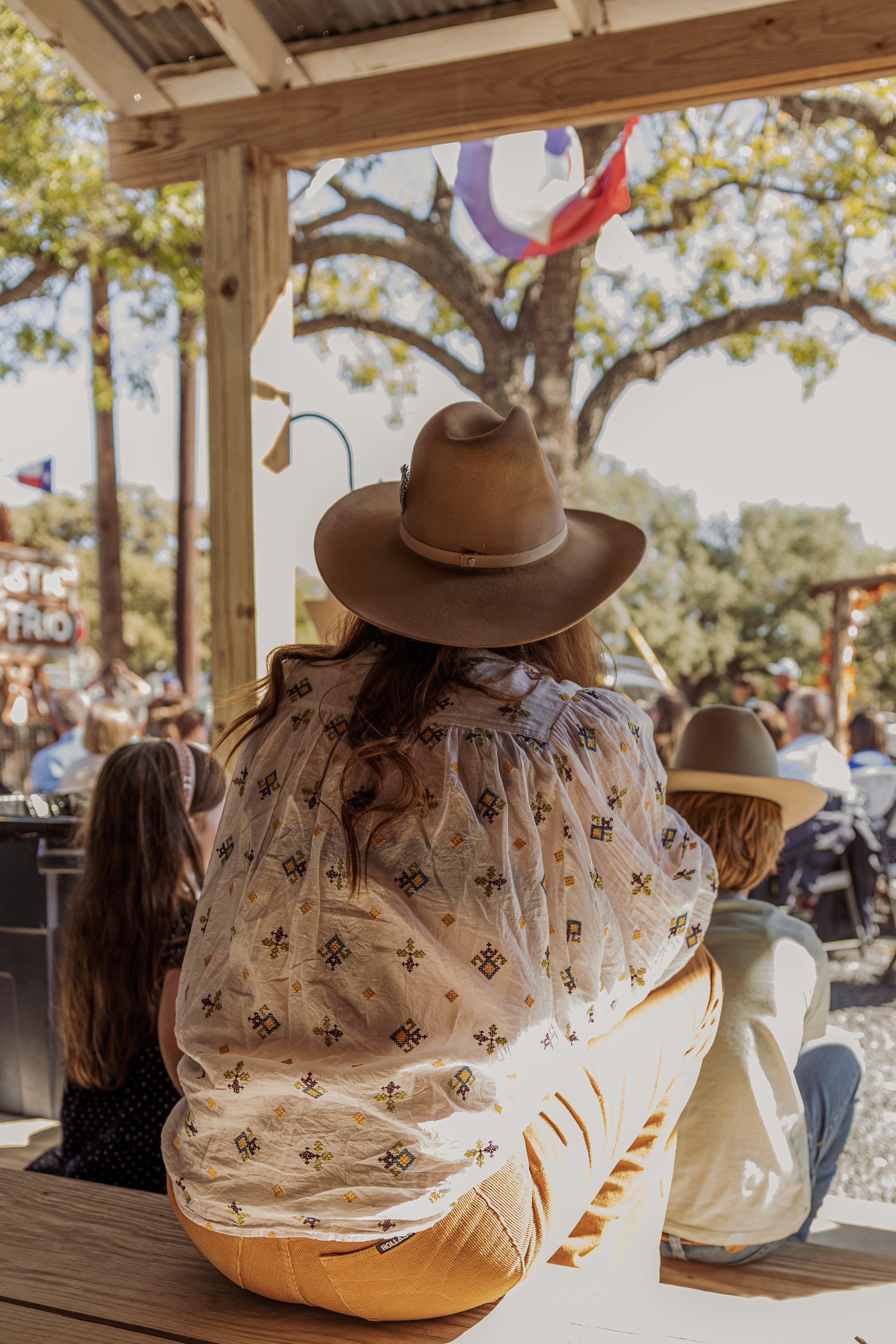 A woman wearing a wide-brimmed tan cowboy hat and a white embroidered shirt sitting on a wooden bench at an outdoor event with a crowd, trees, and hanging flags in the background.