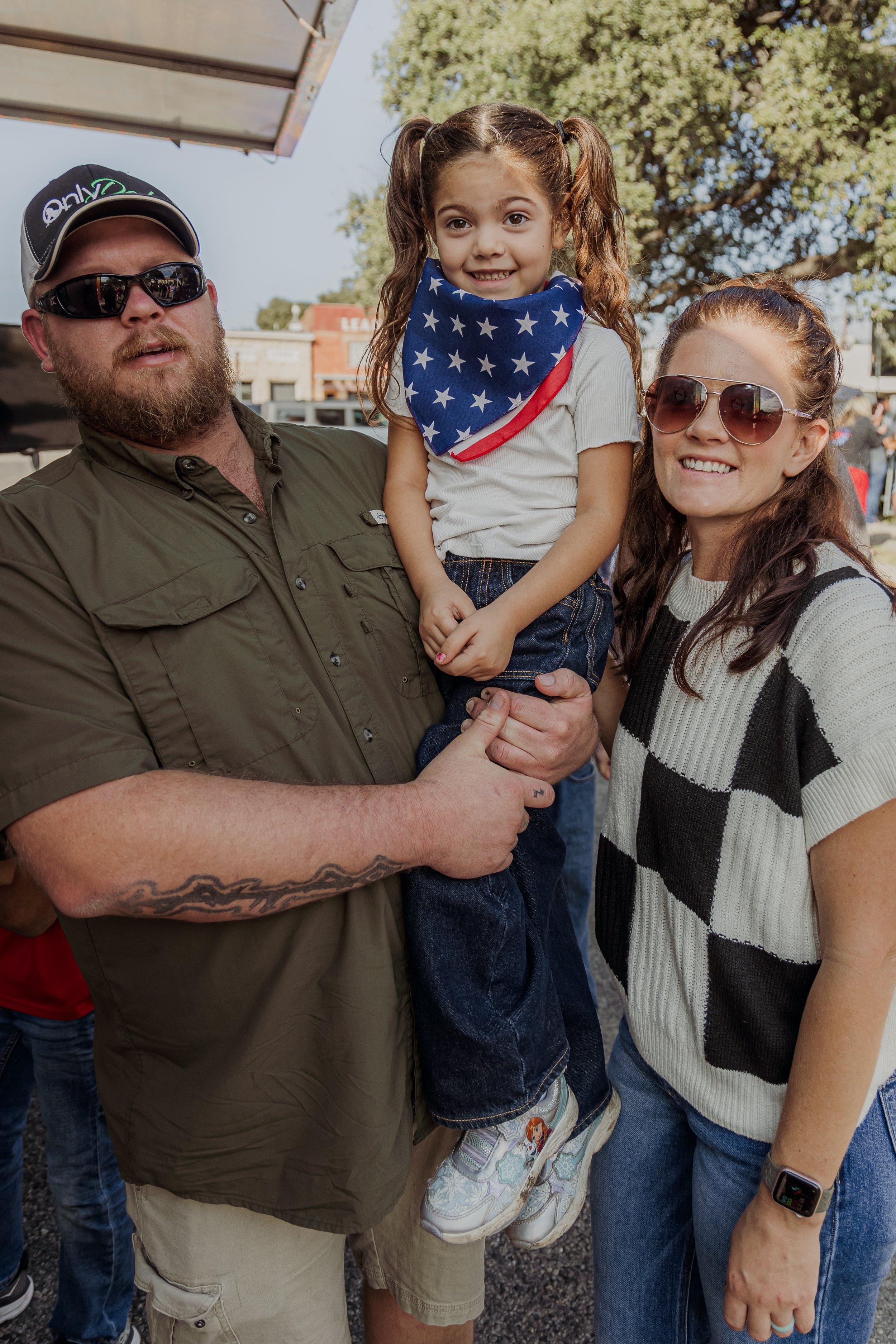 A man holding a young girl with a star-spangled bandana, and a woman standing beside them, outdoors at a festival or fair with trees and buildings in the background.