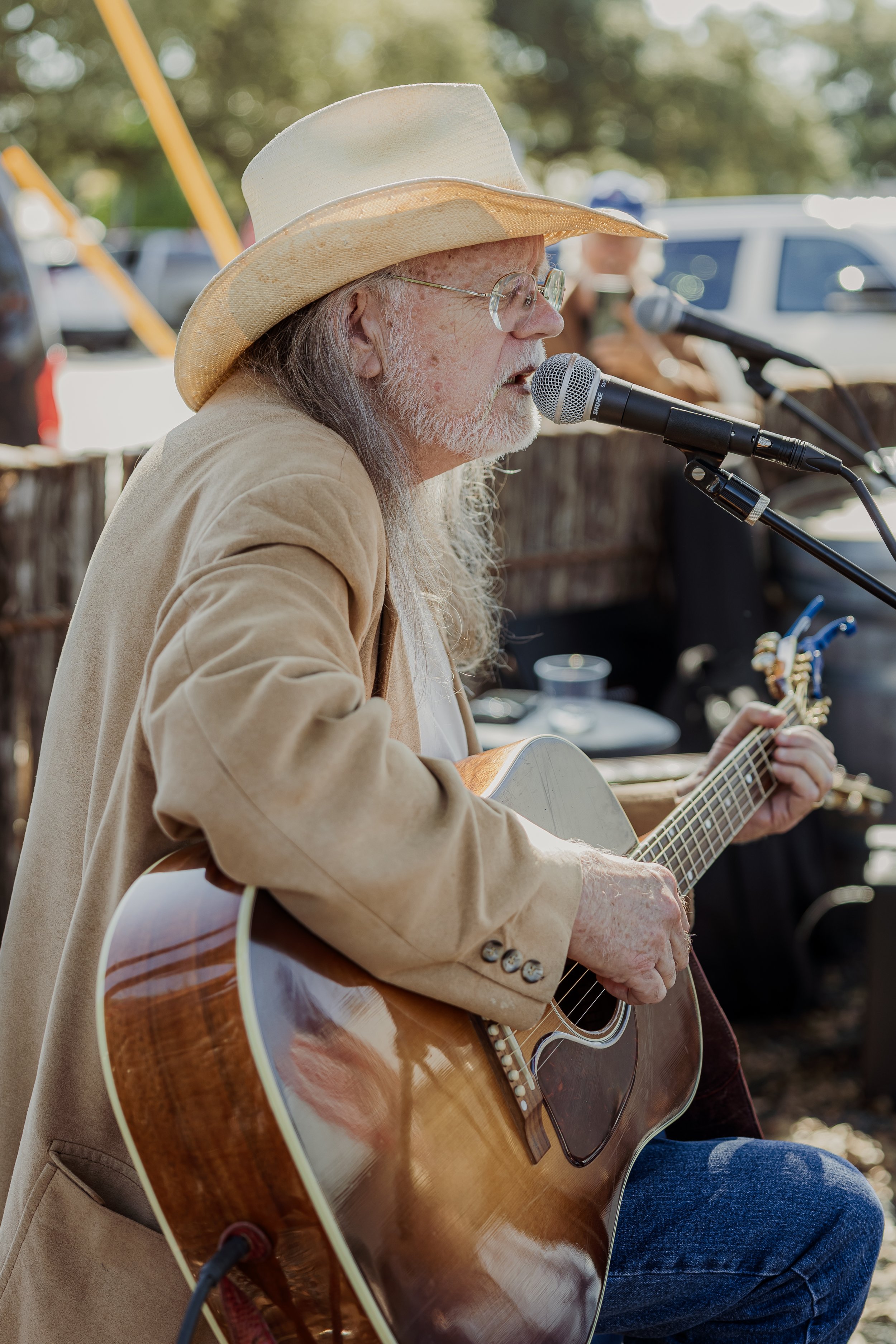 An elderly man with long gray hair, glasses, and a beige cowboy hat playing an acoustic guitar and singing into a microphone at an outdoor event.