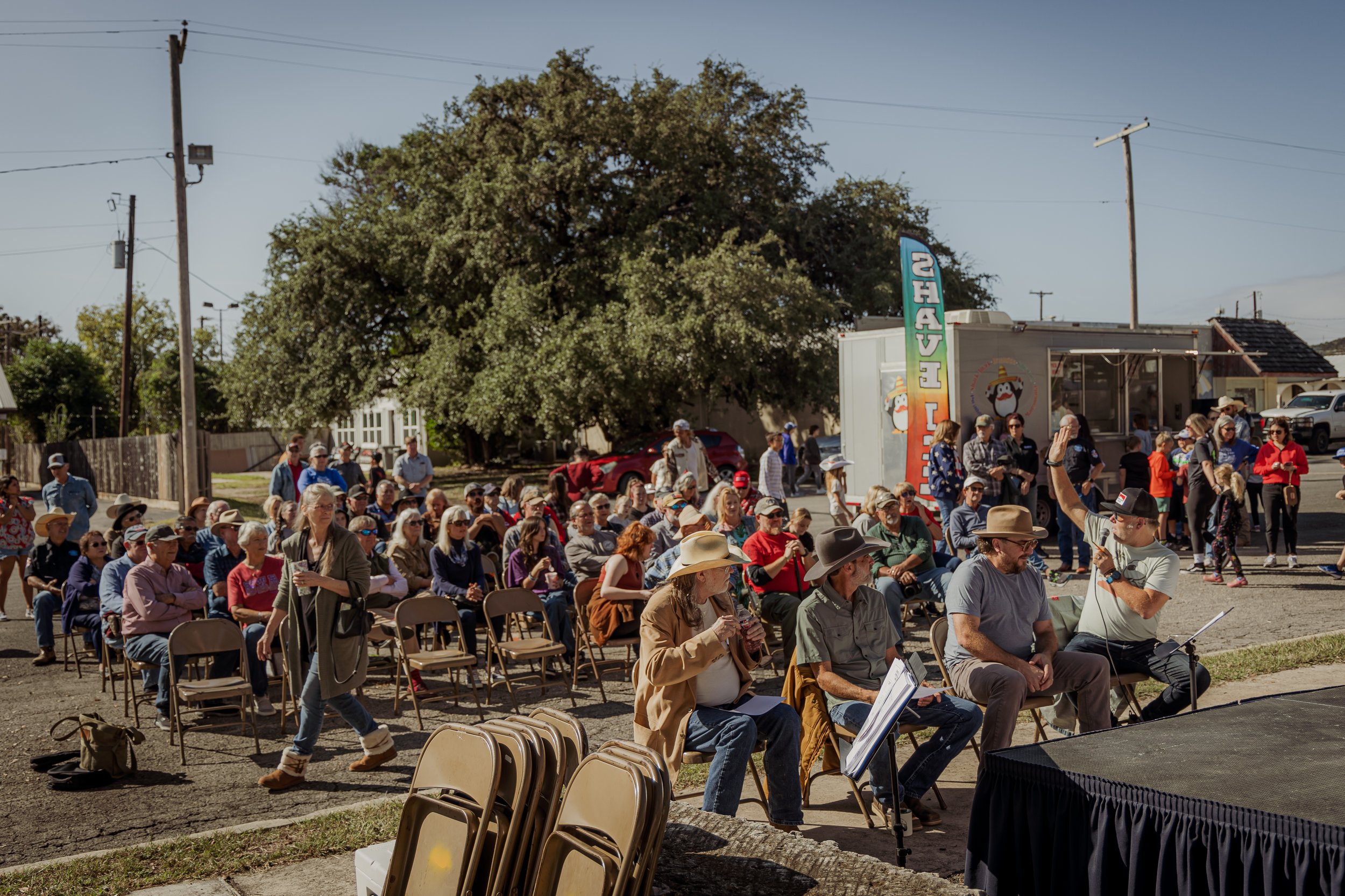 Outdoor event with a large crowd of seated and standing attendees, some wearing cowboy hats, in front of a food truck and a large tree on a sunny day.