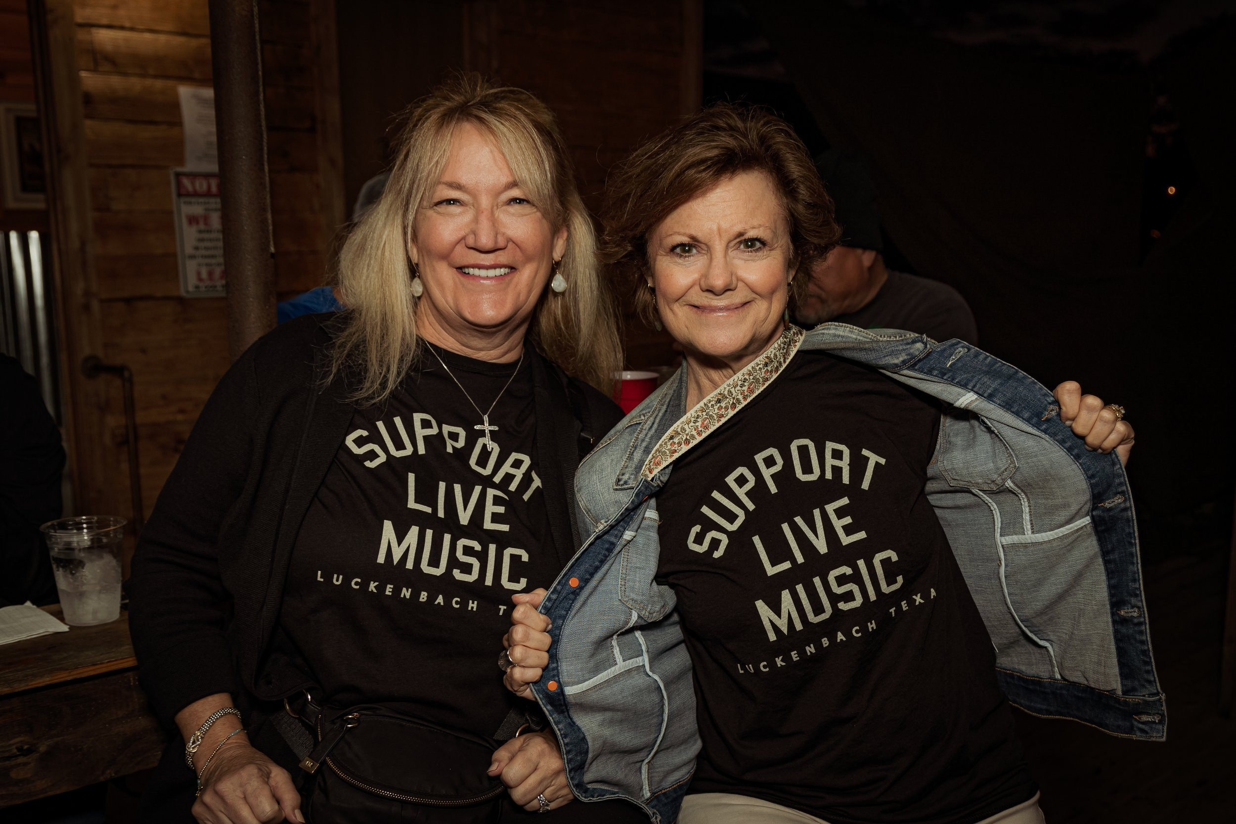 Two women smiling at a music support event, wearing T-shirts with the message 'Support Live Music, Luckenbach Texas.'