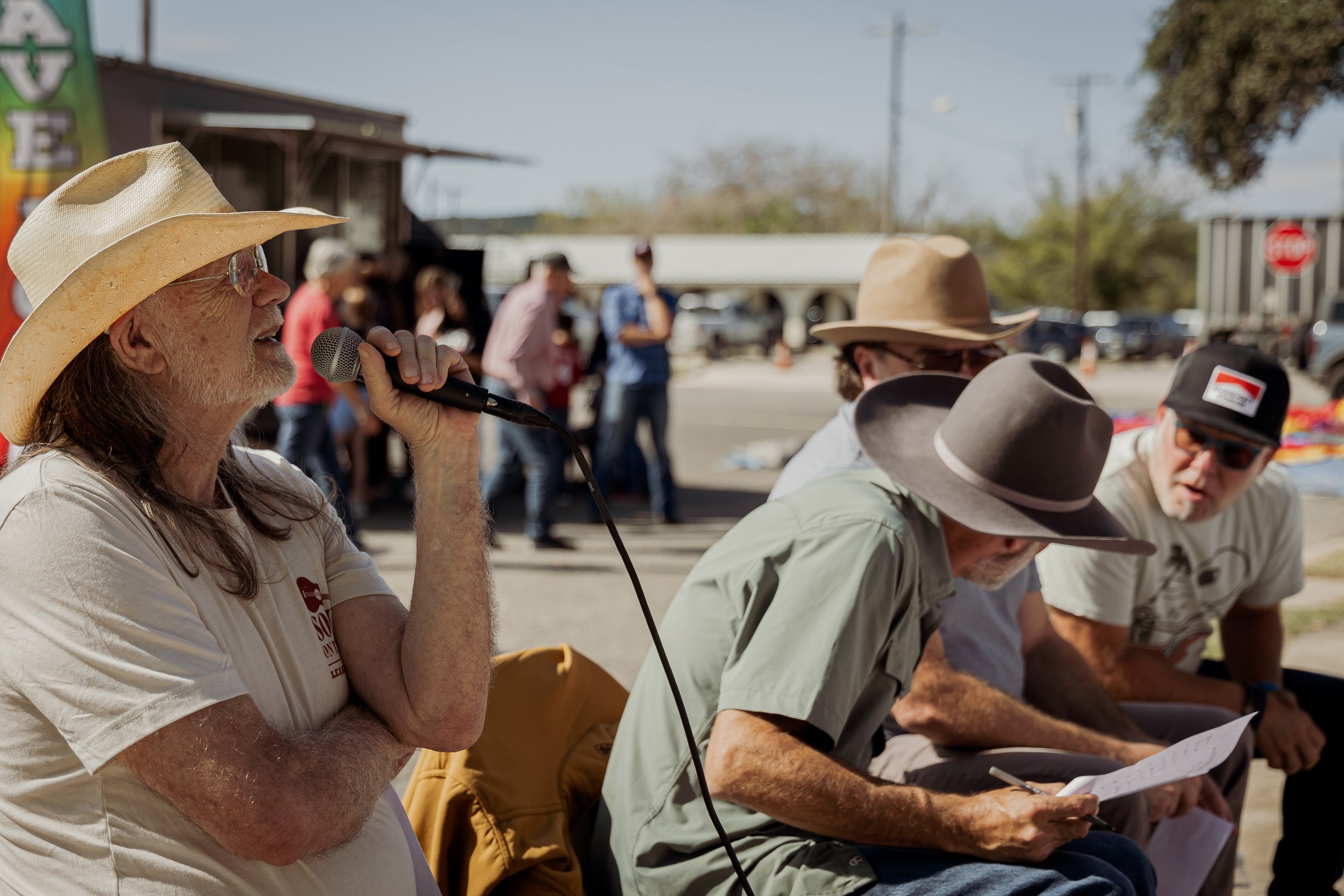 A man wearing a cowboy hat and glasses speaking into a microphone at an outdoor event, with several other men wearing wide-brimmed hats, sitting and taking notes, in a parking lot with people and buildings in the background.