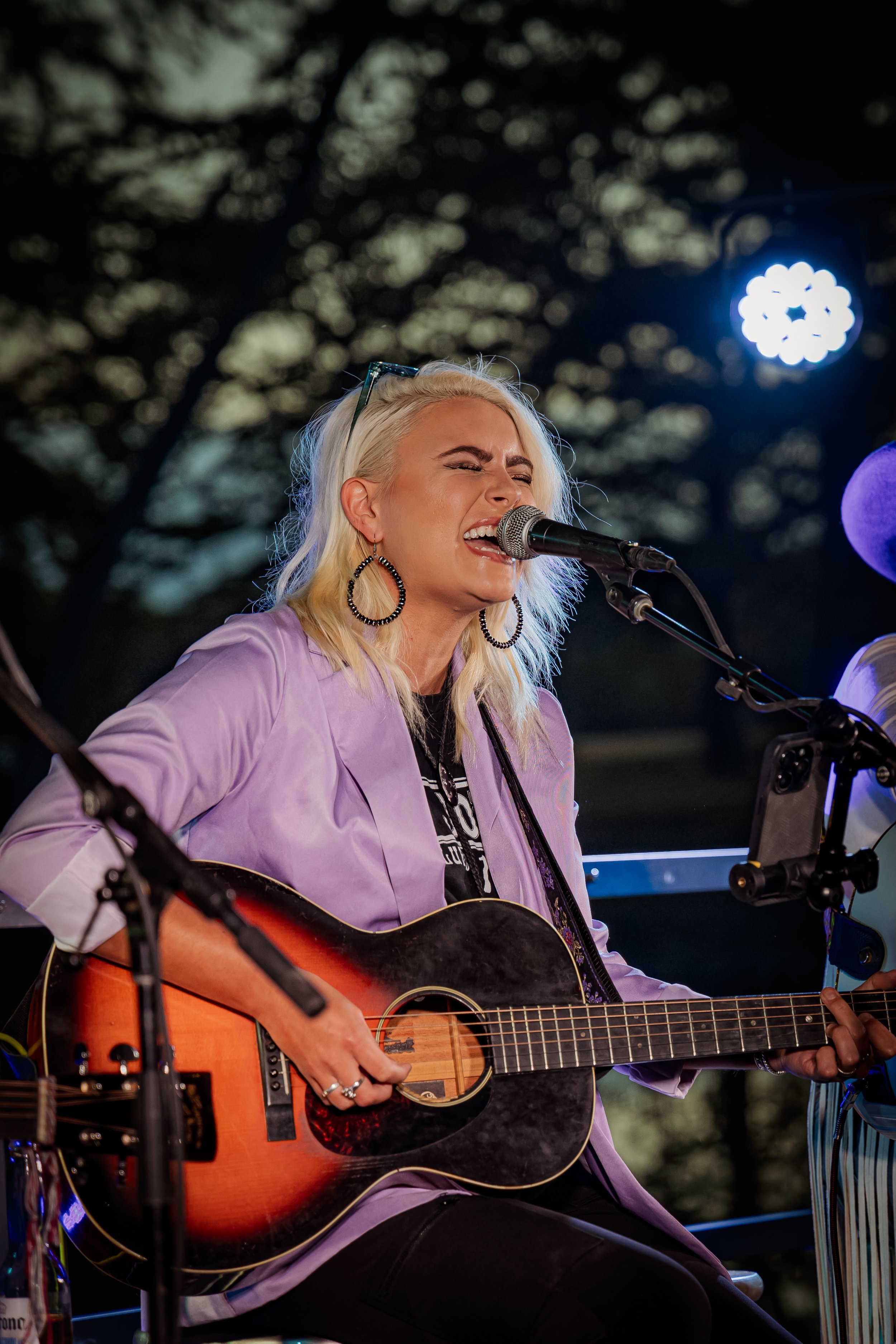 Blonde female musician singing passionately and playing acoustic guitar during outdoor performance at dusk.