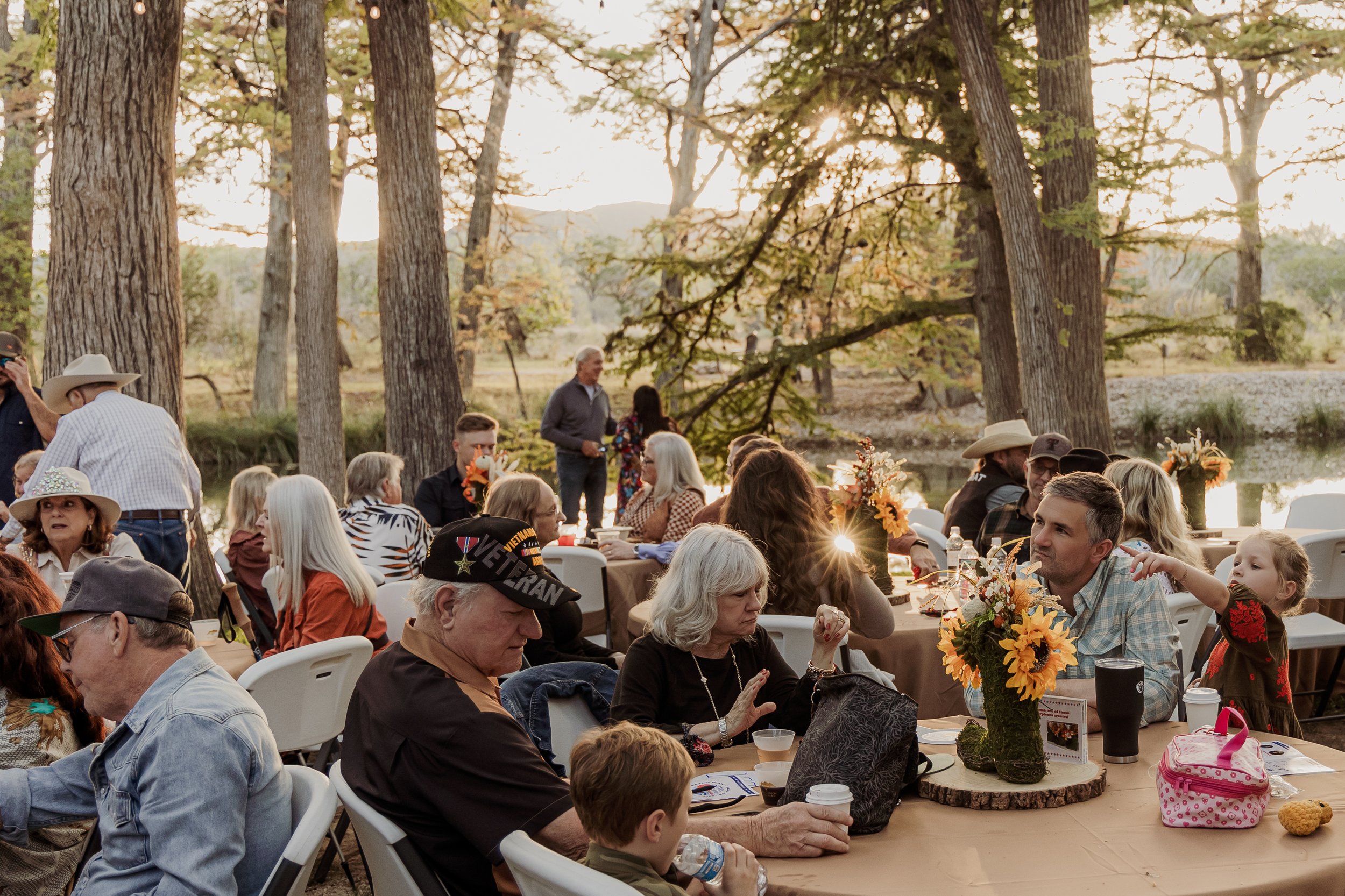 People gathered at an outdoor event during sunset, sitting at tables with floral centerpieces, in a wooded park with tall trees and a pond in the background.