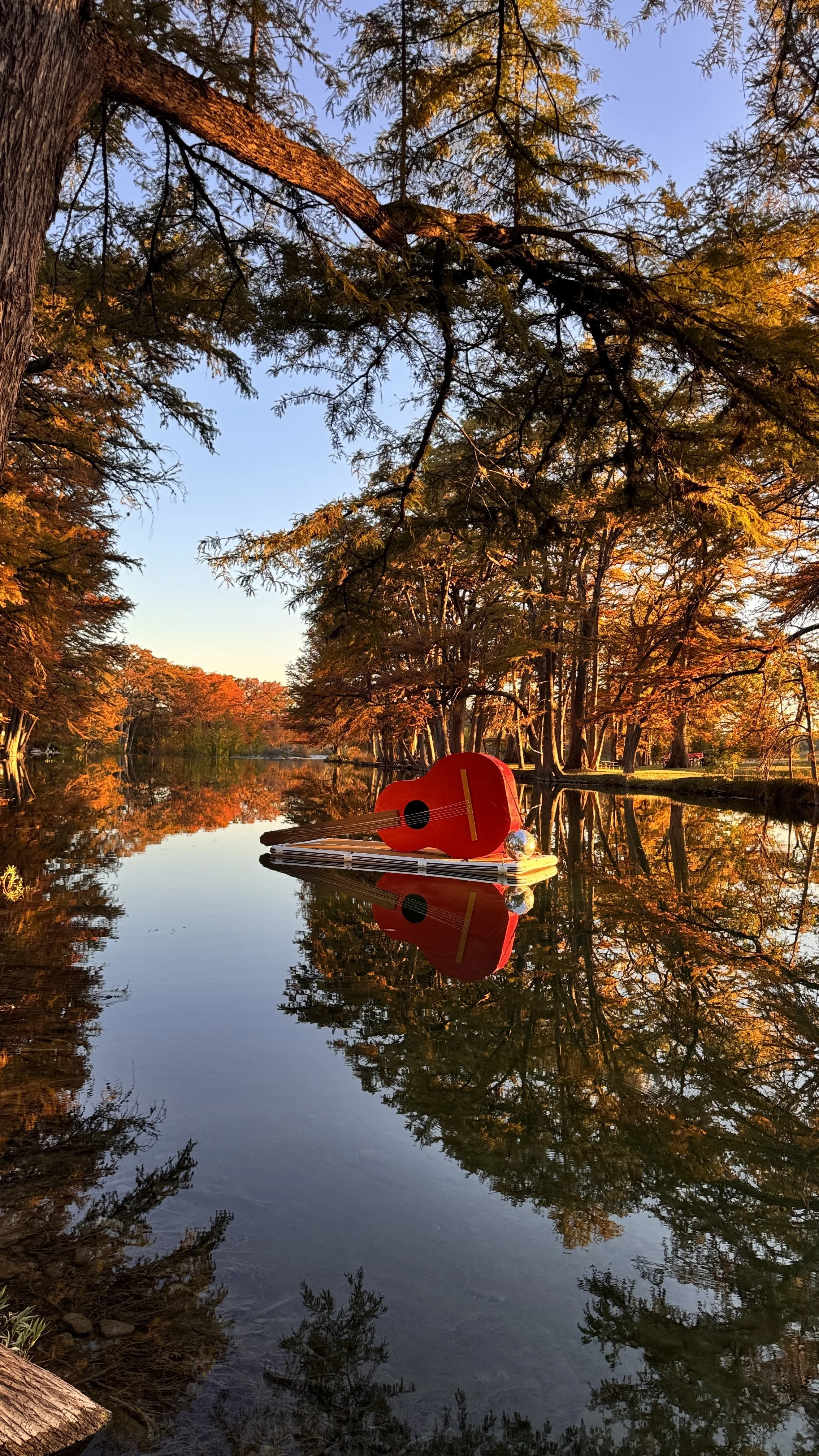A floating dock on a lake with a small red guitar, a shiny silver ball, and a white object in the water. Autumn trees with orange and yellow leaves surround the lake, reflected in the water, under a clear blue sky.